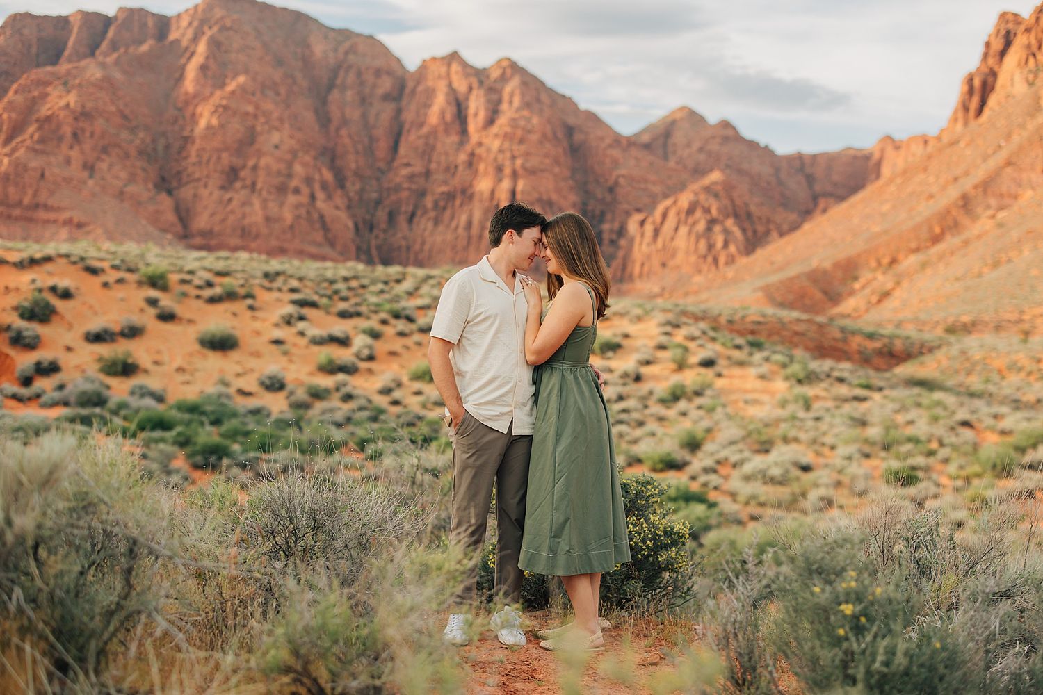 Couple embracing and kissing in desert landscape with red rock mountains and sagebrush at golden hour.