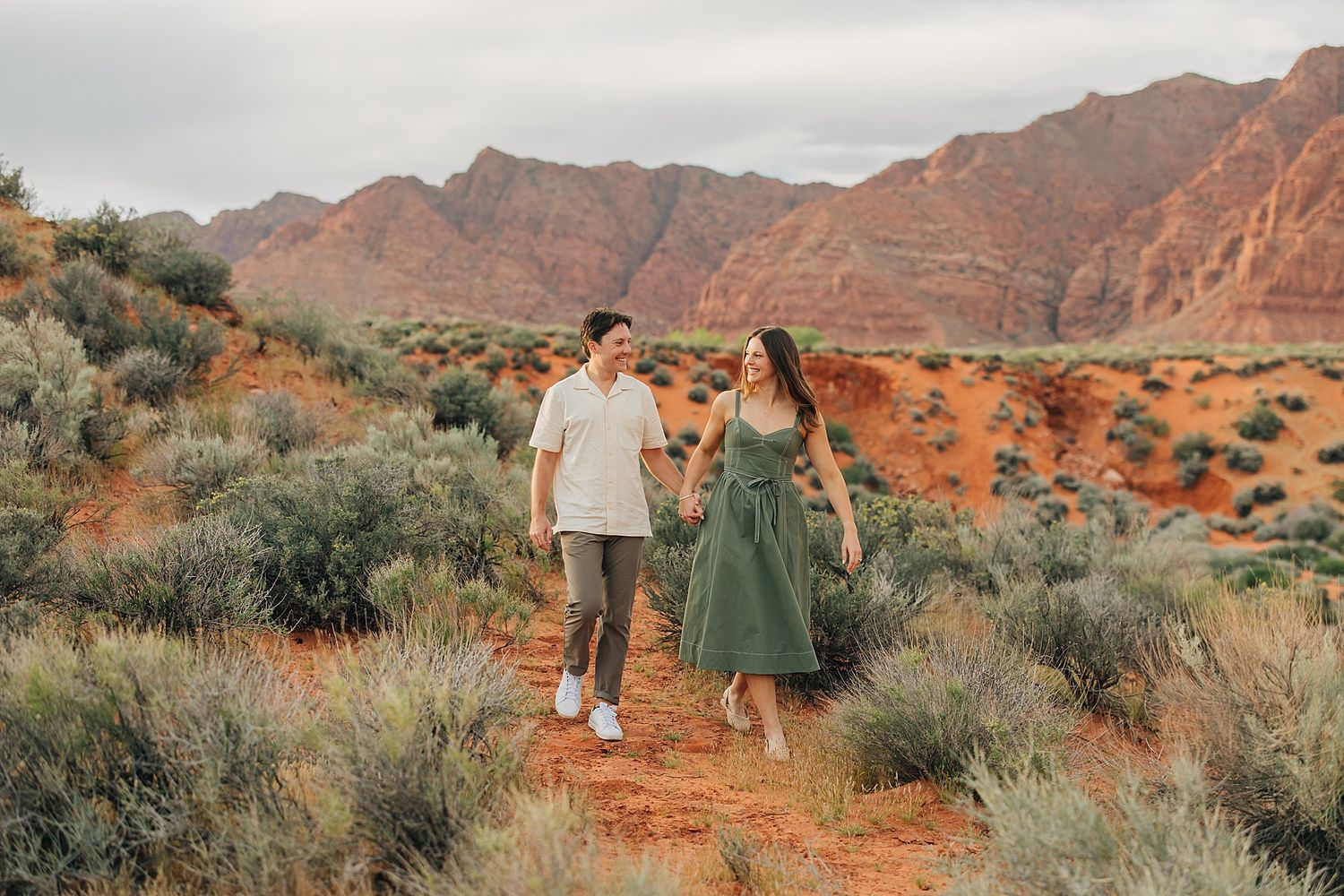 Couple holding hands walking on a red dirt trail surrounded by desert shrubs with red rock mountains in the background.