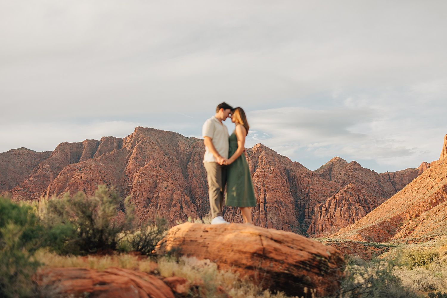 Couple embracing on red rock formation with stunning desert mountain backdrop at golden hour.