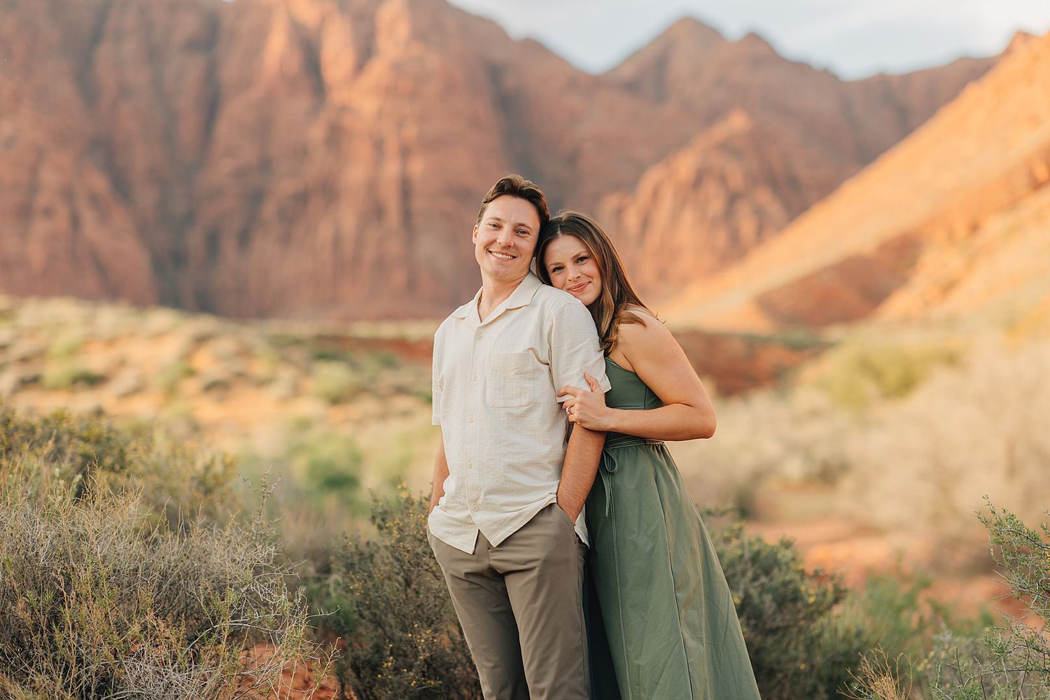Couple posing together in a desert landscape with red rock mountains, man in white shirt and woman in green dress.
