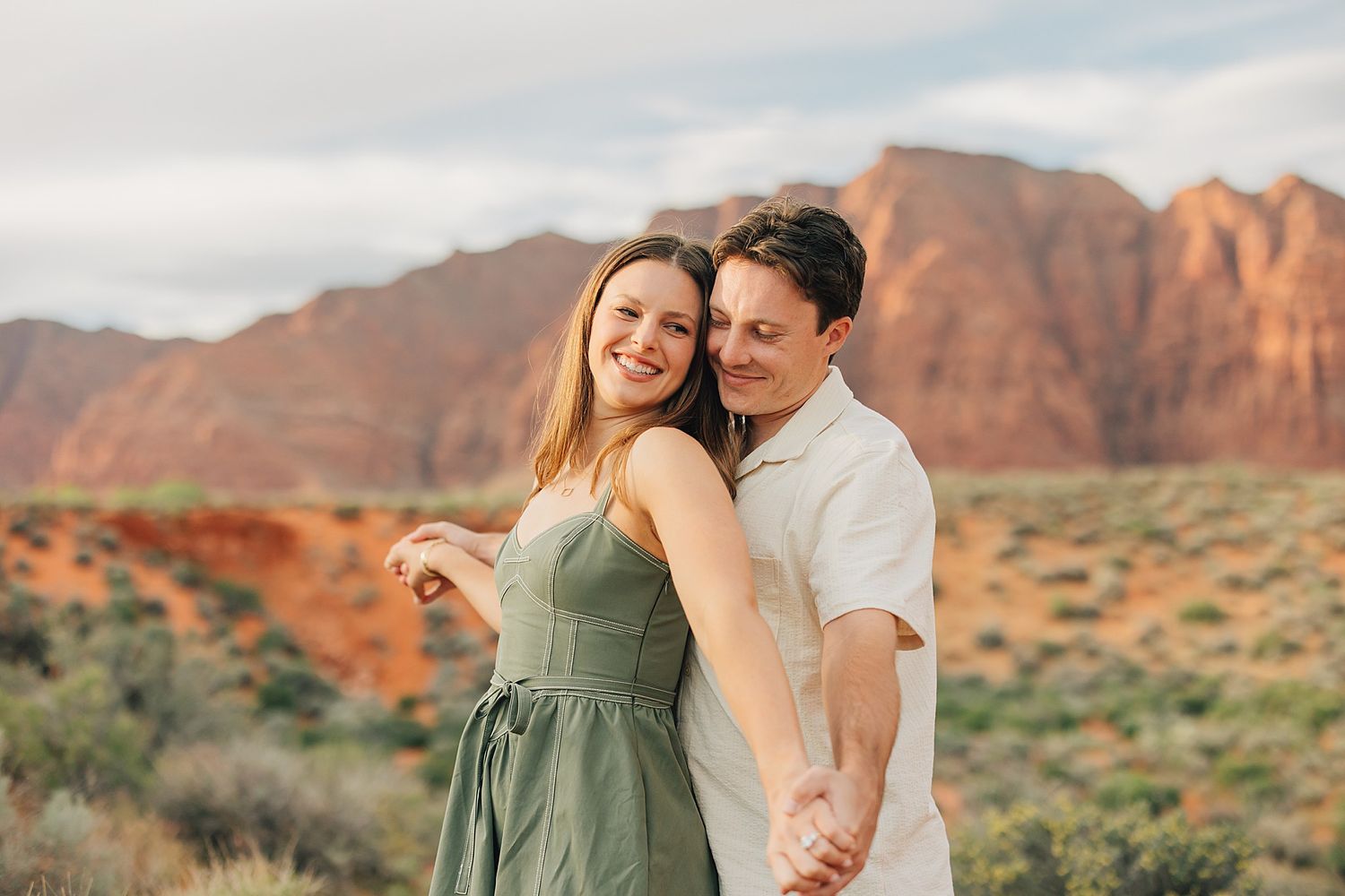 Smiling couple holding hands in front of red rock canyon landscape wearing green dress and white shirt.