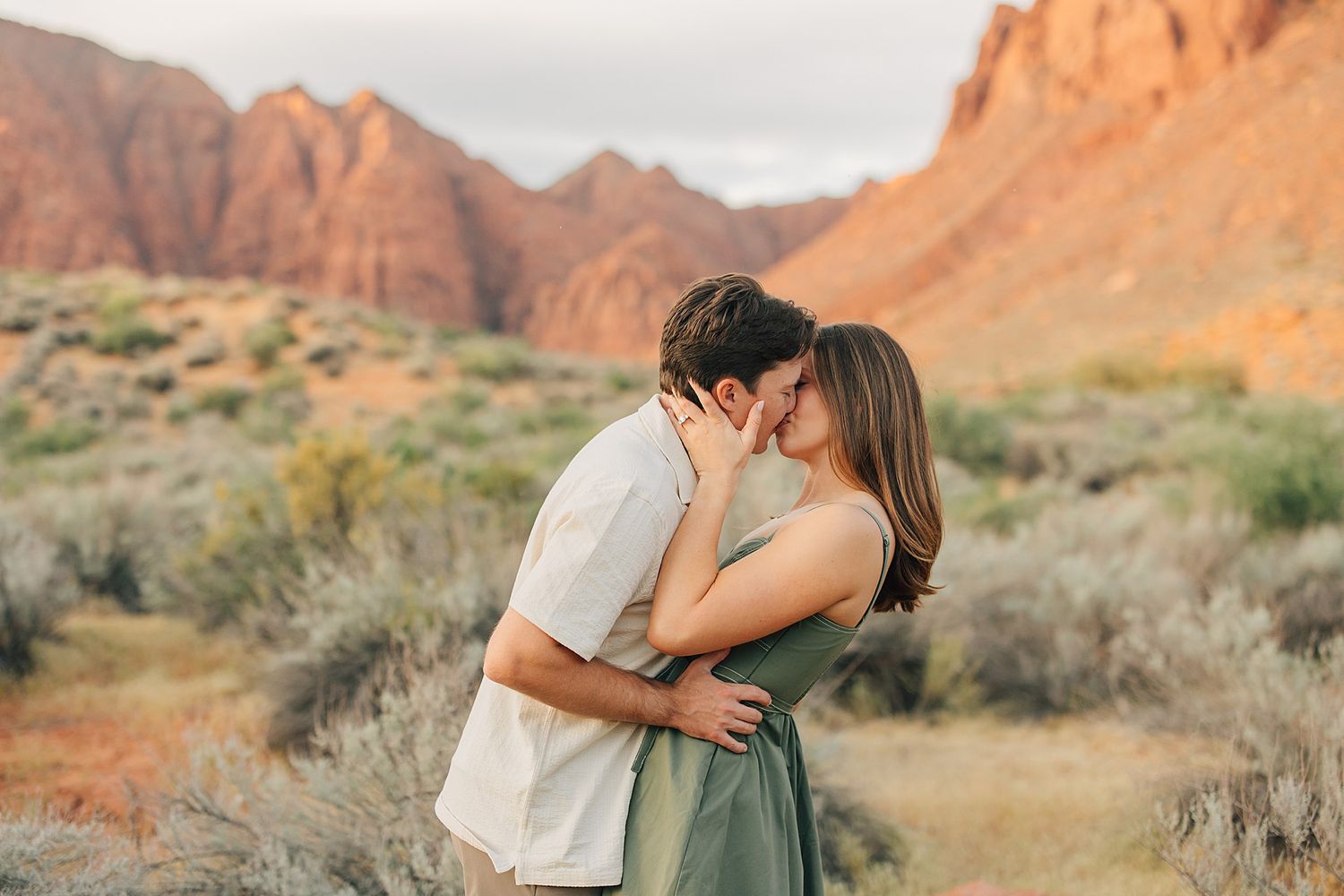 Couple sharing a romantic kiss surrounded by desert shrubs and red sandstone cliffs at sunset.