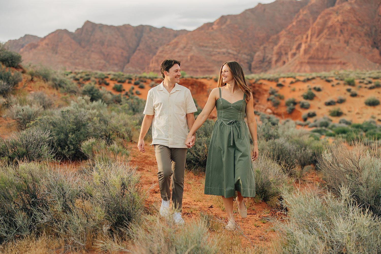 Couple holding hands walking through red desert landscape with sandstone cliffs and sage brush in background.