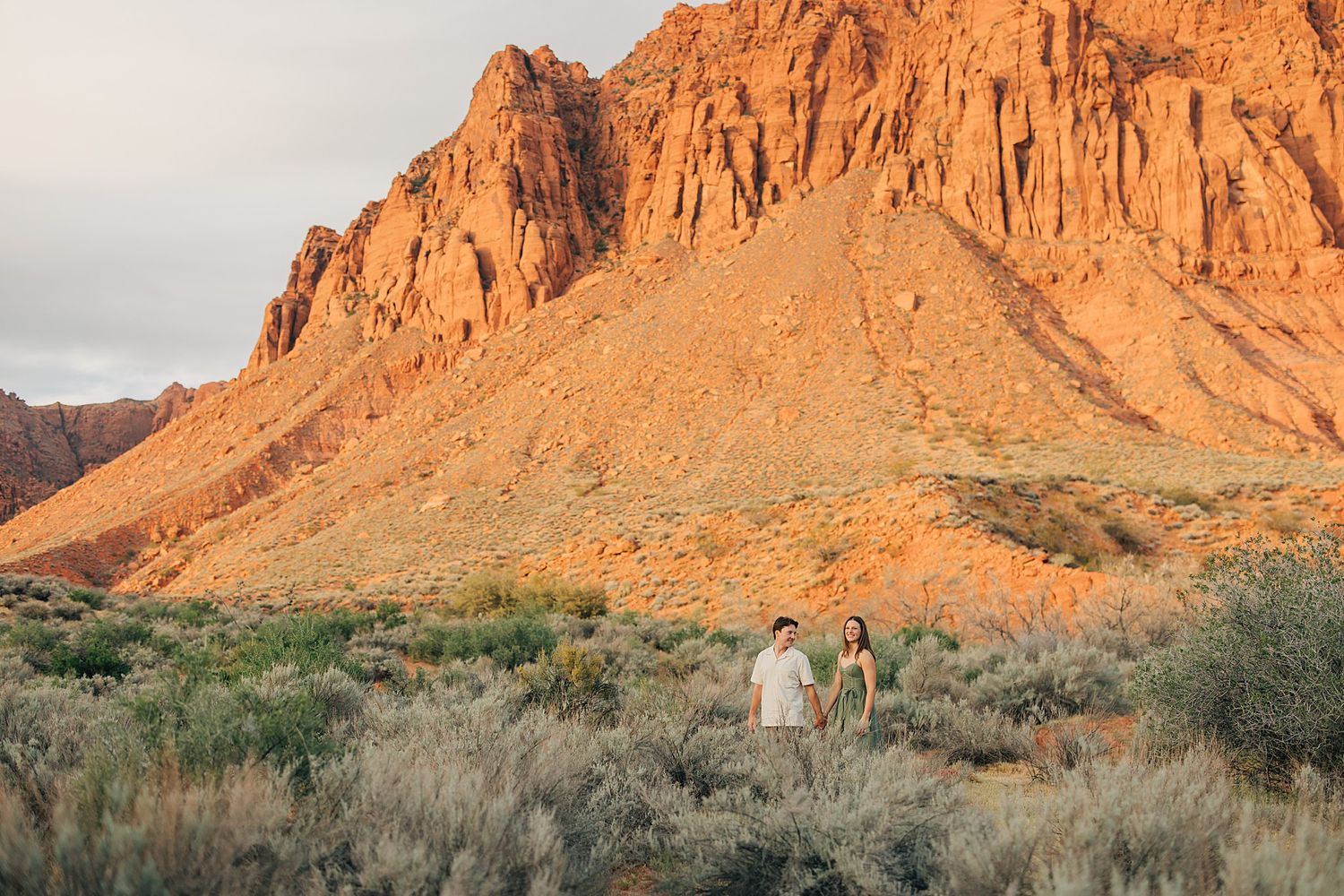 Couple holds hands in desert landscape with dramatic red rock mountain backdrop at golden hour.