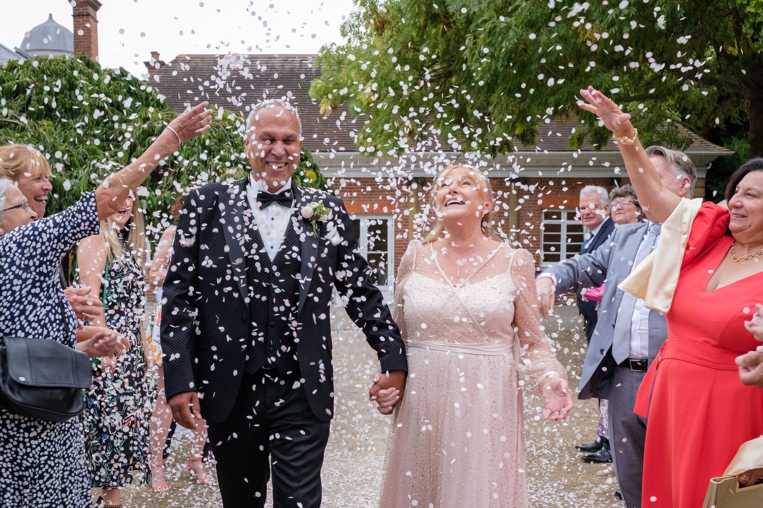 Joyful couple walks through confetti shower at outdoor wedding celebration with guests cheering around them.