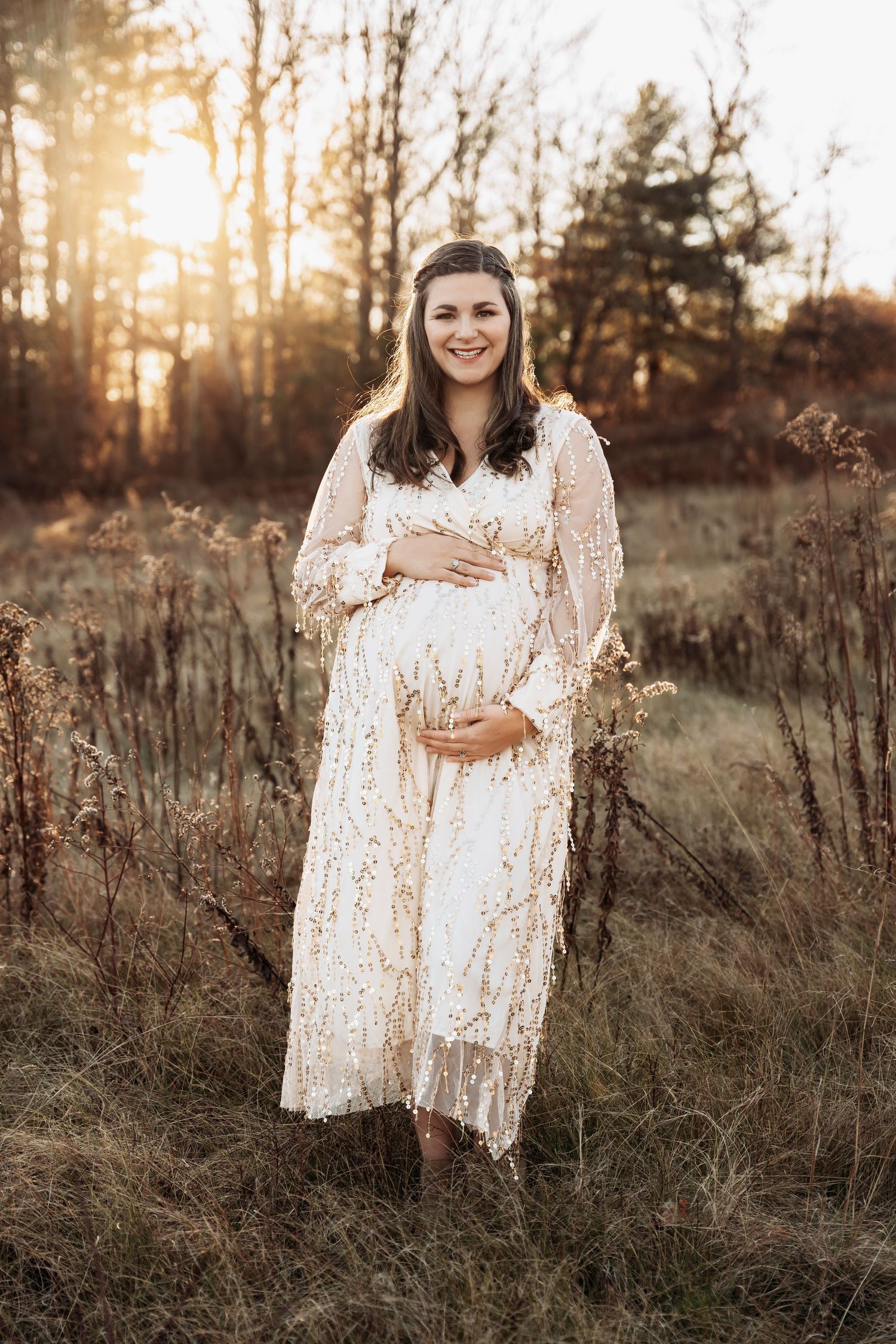 Smiling pregnant woman in white lace dress cradling baby bump in golden sunset field.
