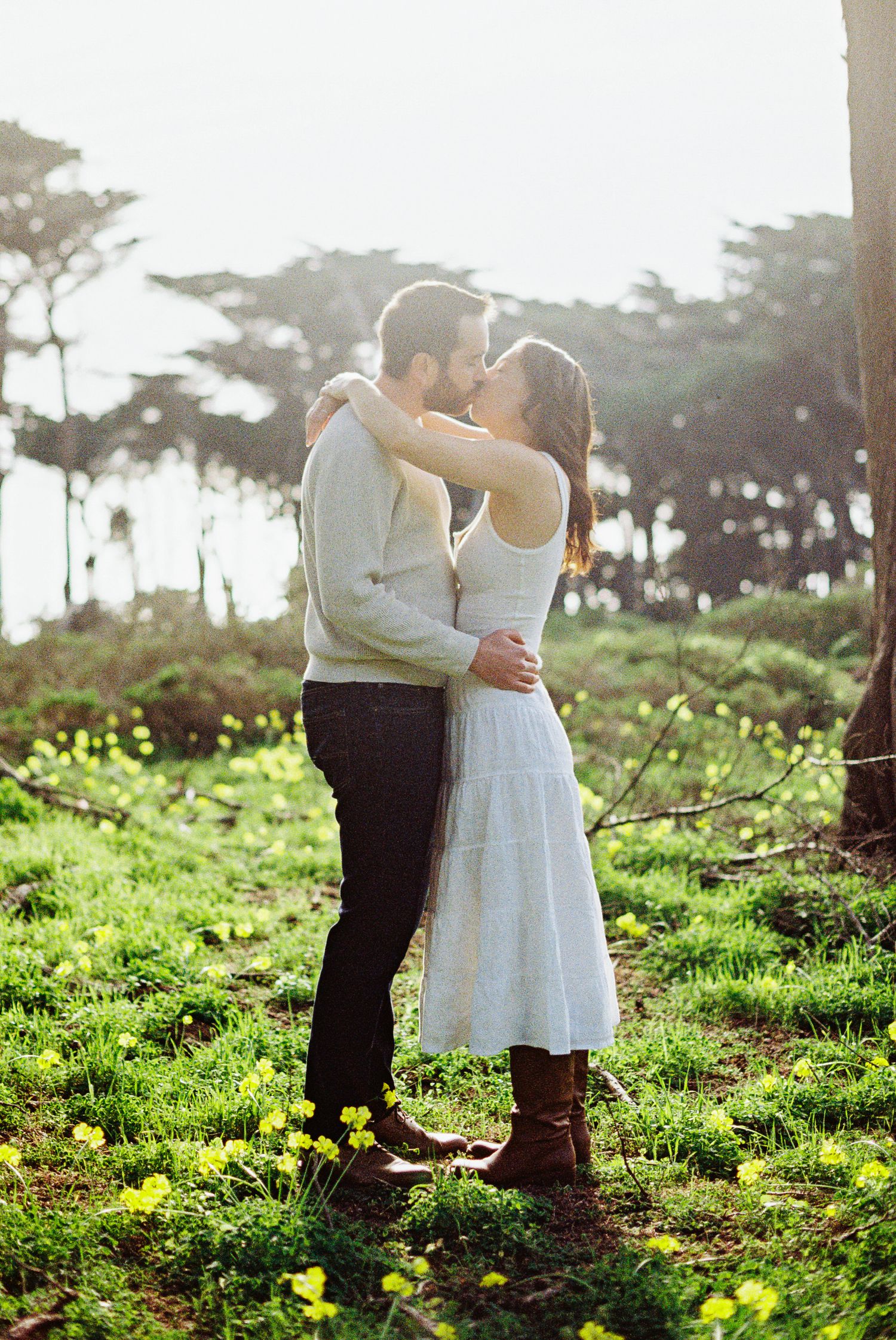 Couple sharing a romantic kiss in a sunlit forest meadow with yellow wildflowers, woman in white dress and brown boots.