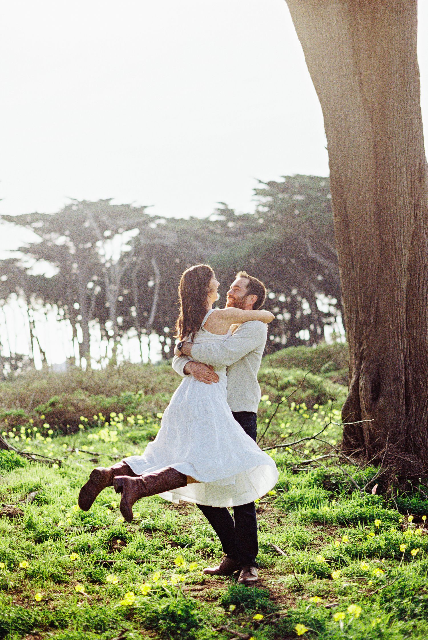 A couple embraces and spins joyfully in a lush green meadow surrounded by tall trees in soft natural light.