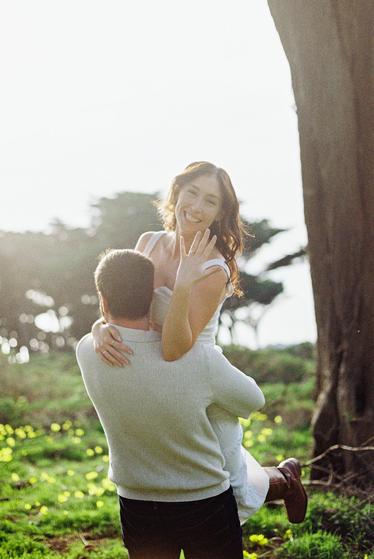 A joyful woman shows off her engagement ring while being lifted by her partner outdoors.