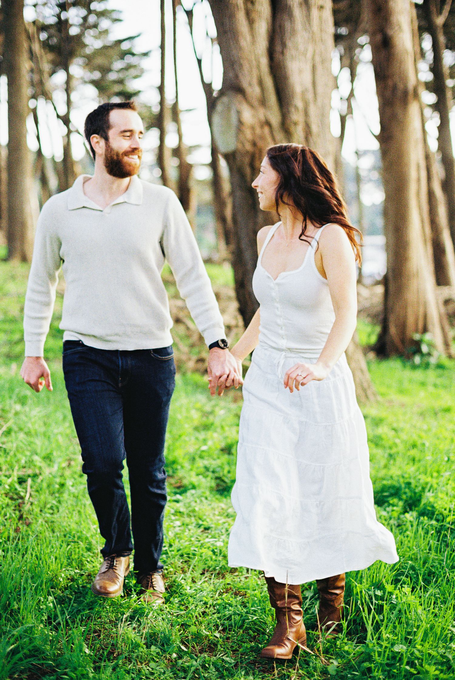 Couple holding hands and smiling at each other while walking through a lush green forest clearing.