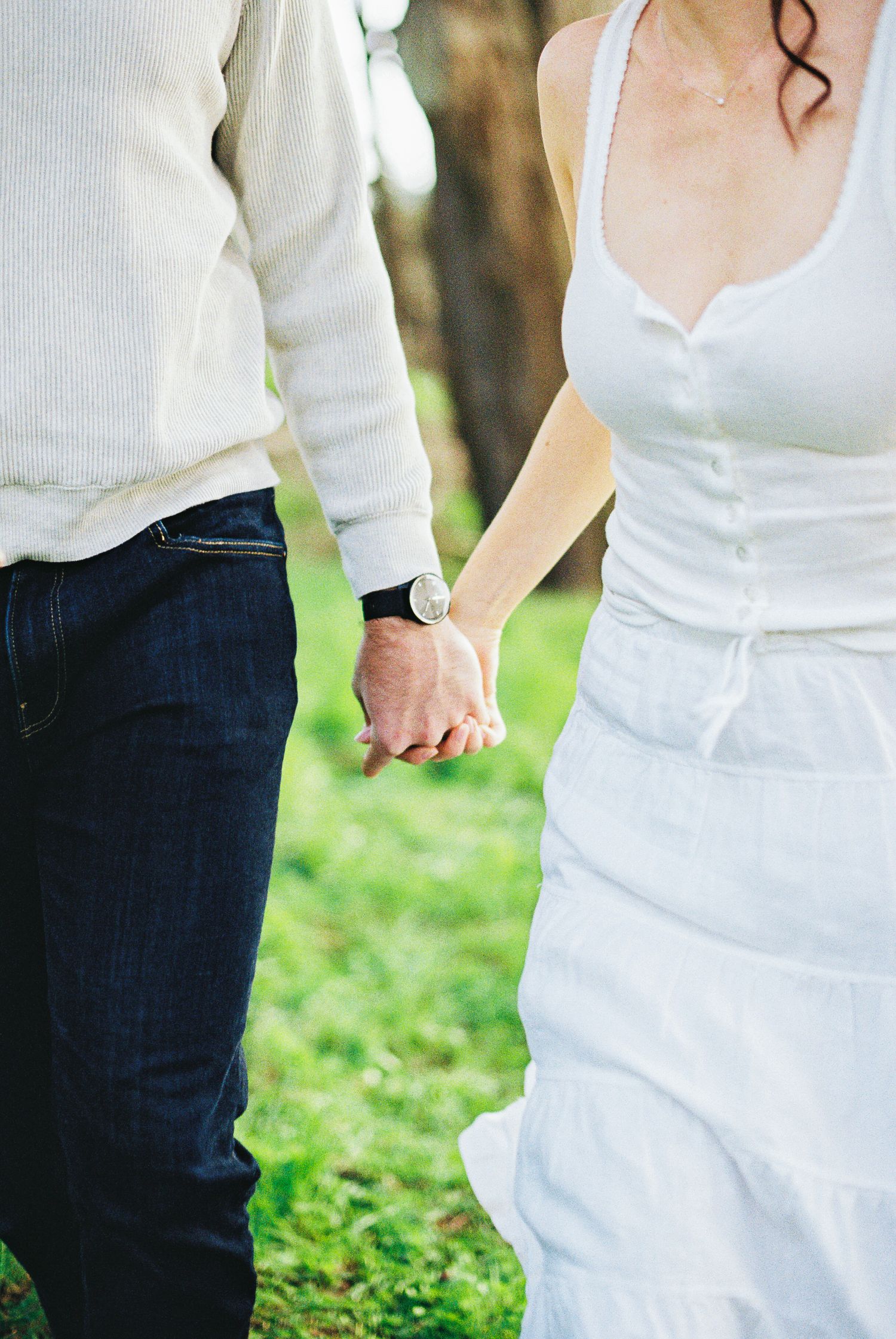 Couple holding hands outdoors in a garden, man in jeans and watch, woman in white dress on green grass.