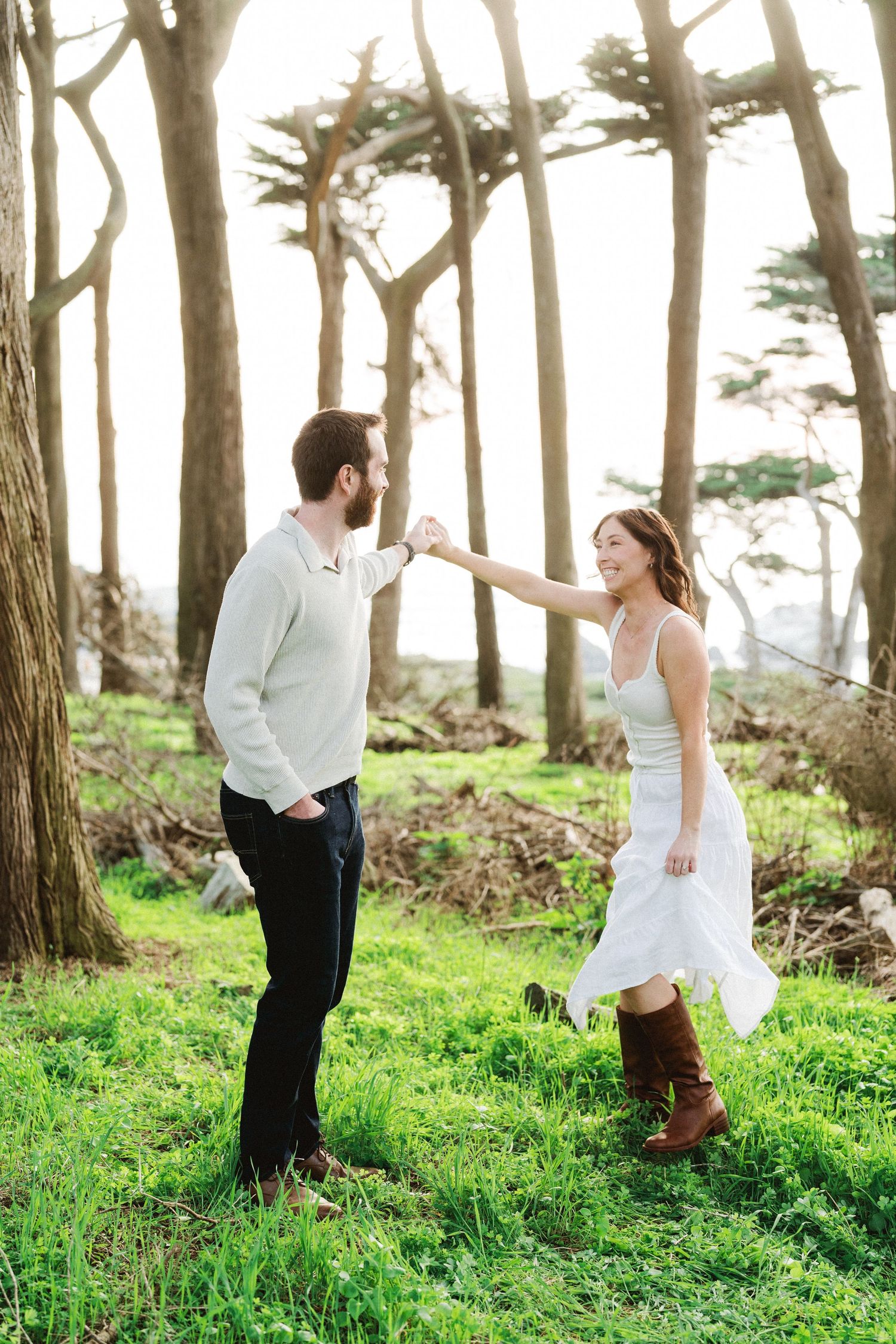 Couple dancing joyfully in a sunlit forest clearing with lush green grass and tall trees.