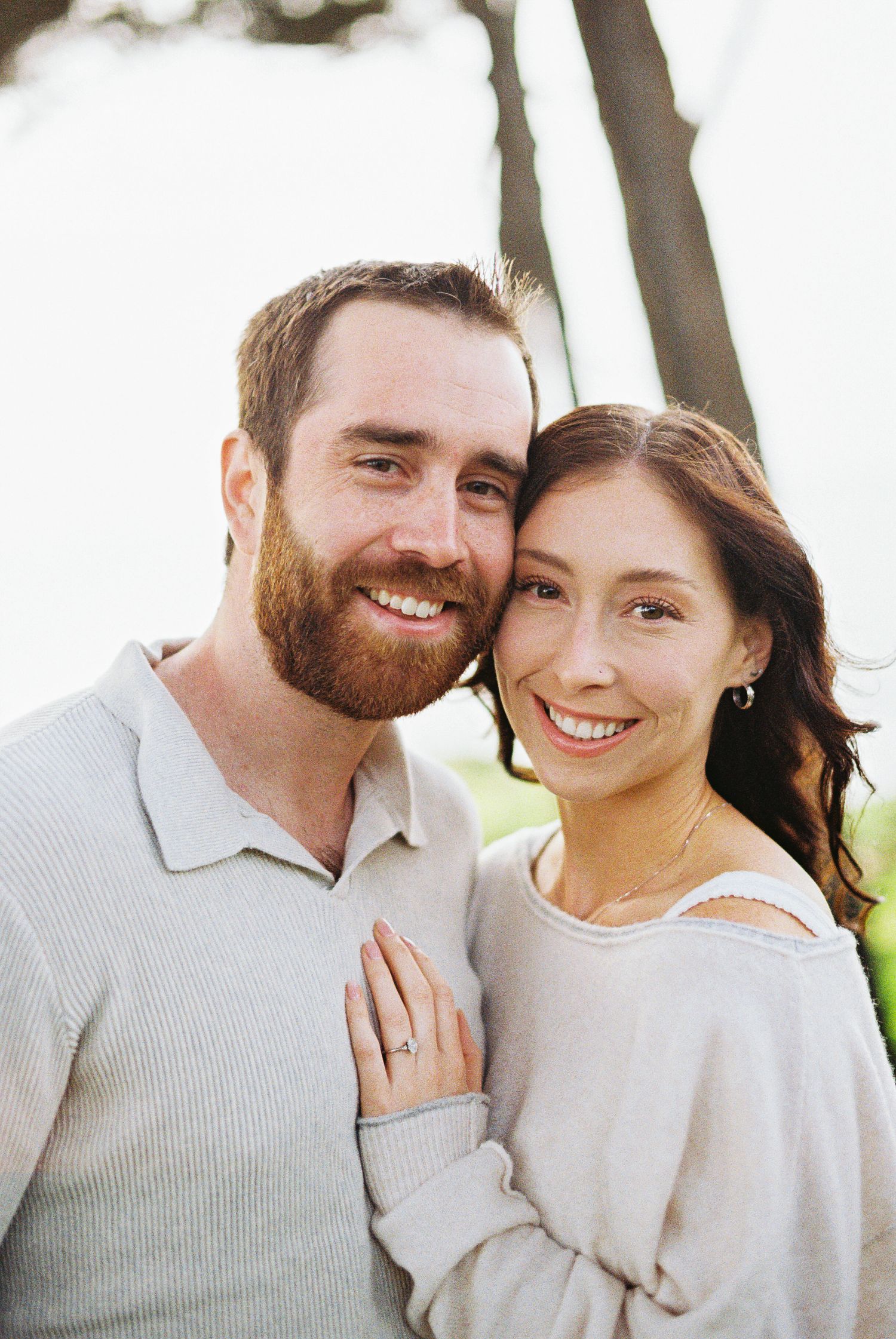 Smiling couple posing outdoors, woman showing engagement ring, man with red beard, both wearing casual light-colored sweaters.