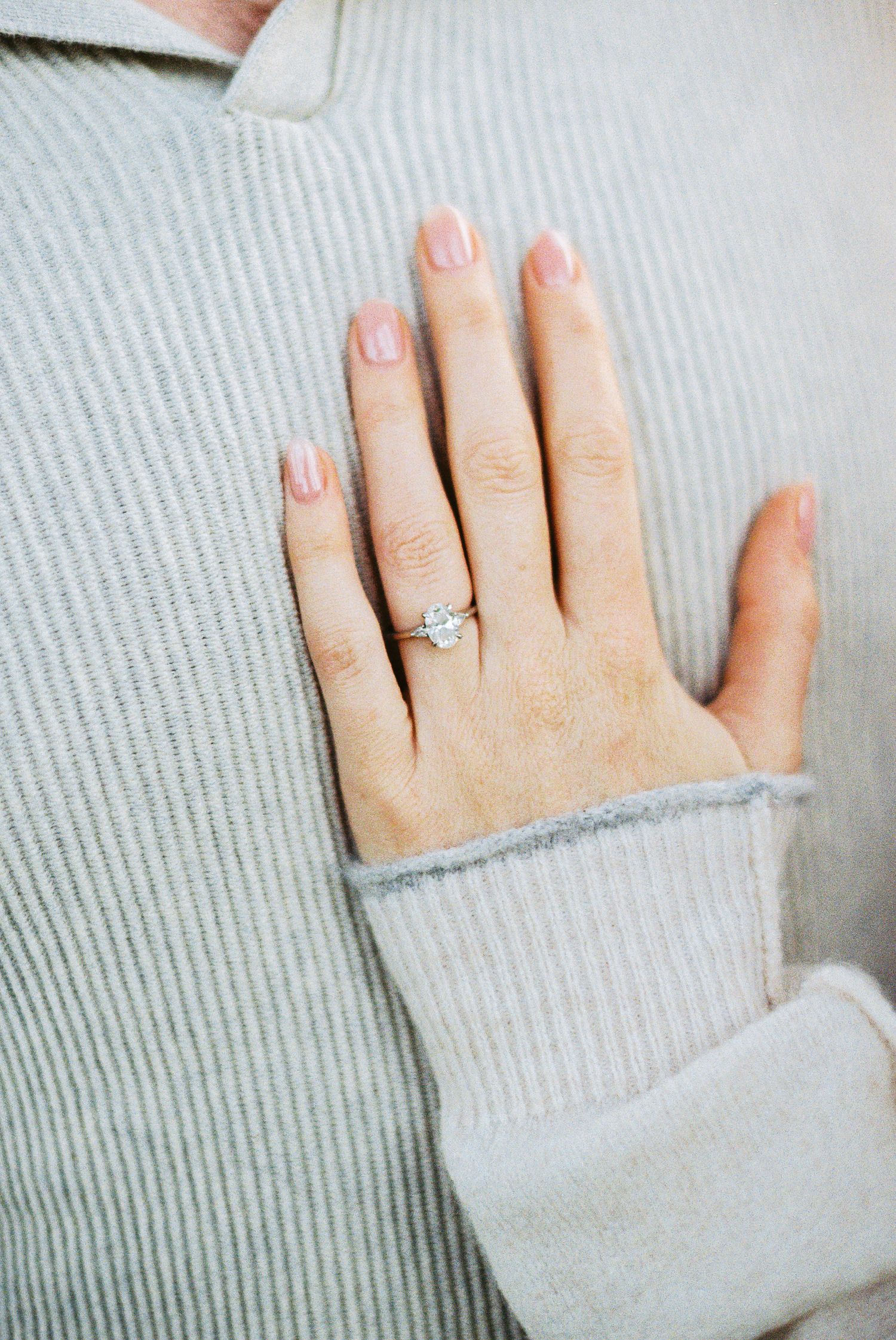 Close-up of a hand wearing a solitaire diamond engagement ring with pink manicure on a gray ribbed sweater.