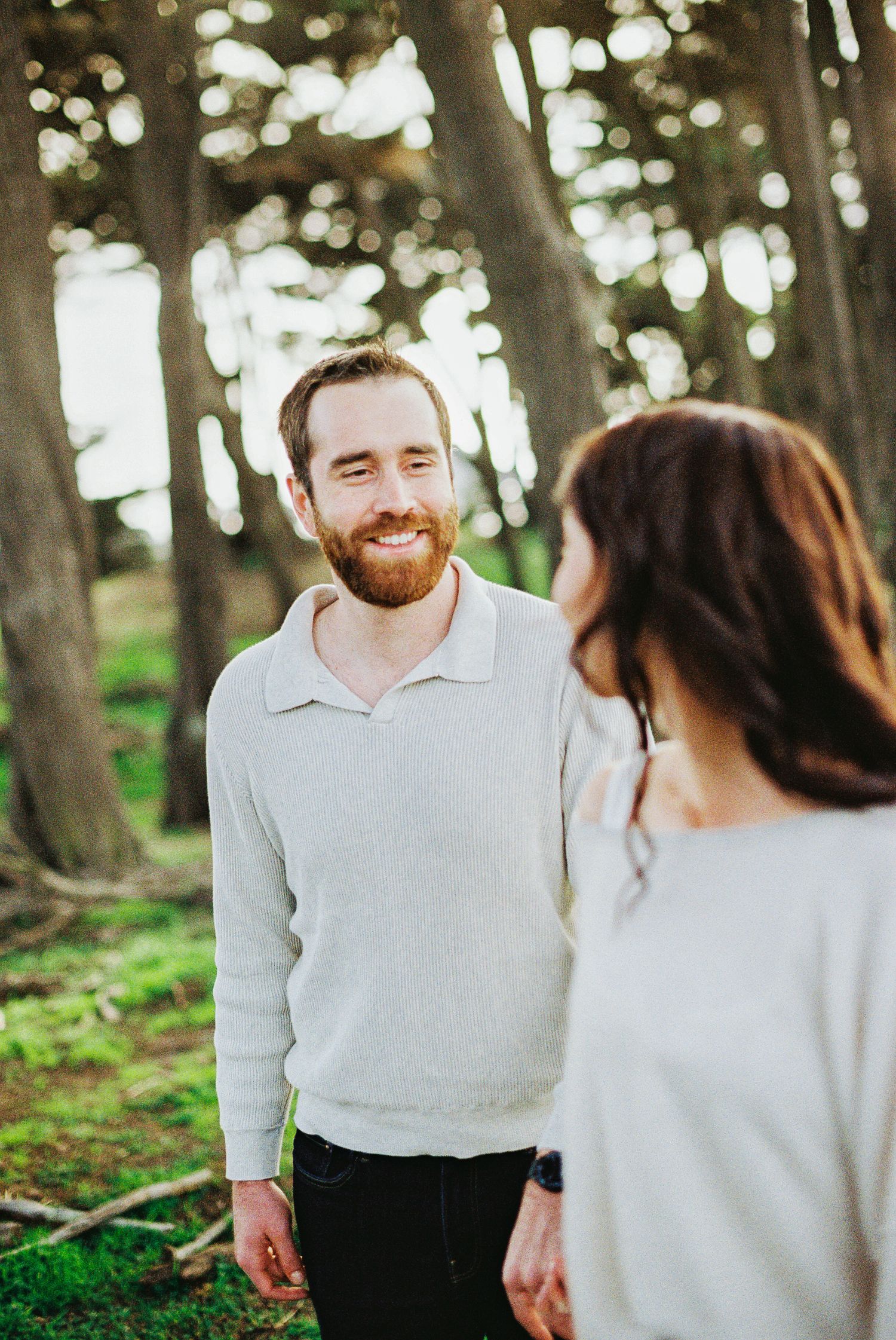 A smiling bearded man in a white sweater gazes at a woman outdoors in a lush green forest setting.