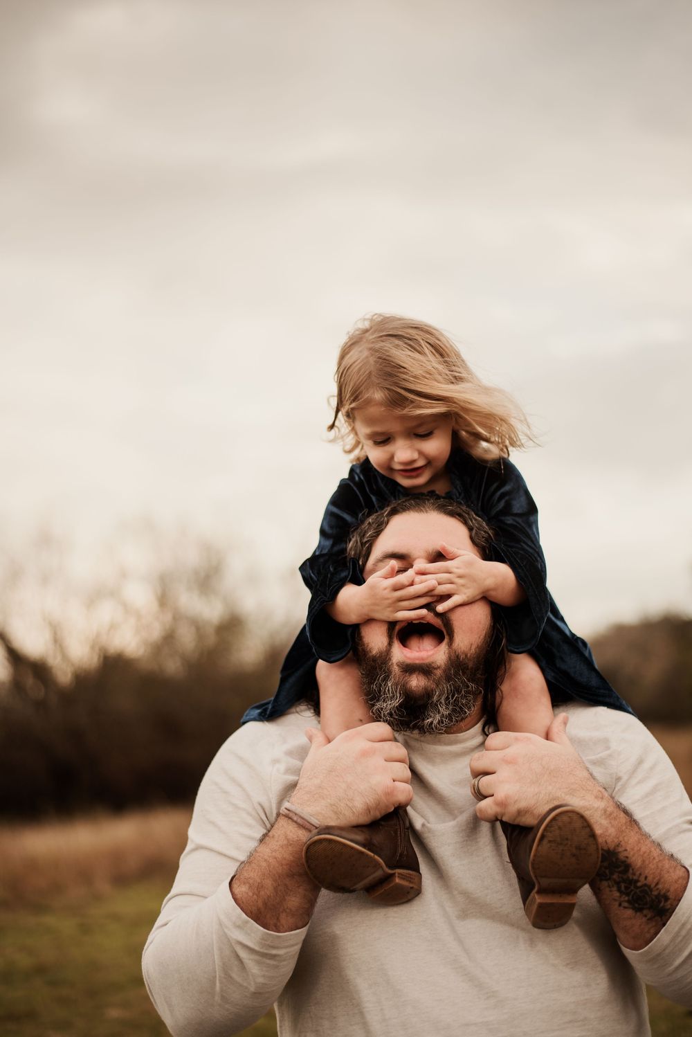 A bearded man smiles as a young girl sits on his shoulders covering his eyes outdoors.