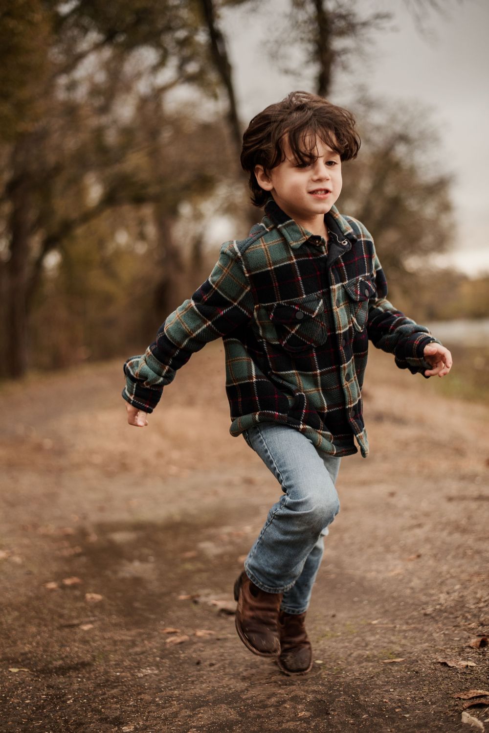 A young boy in a plaid flannel shirt and jeans runs joyfully down a dirt path surrounded by autumn trees.