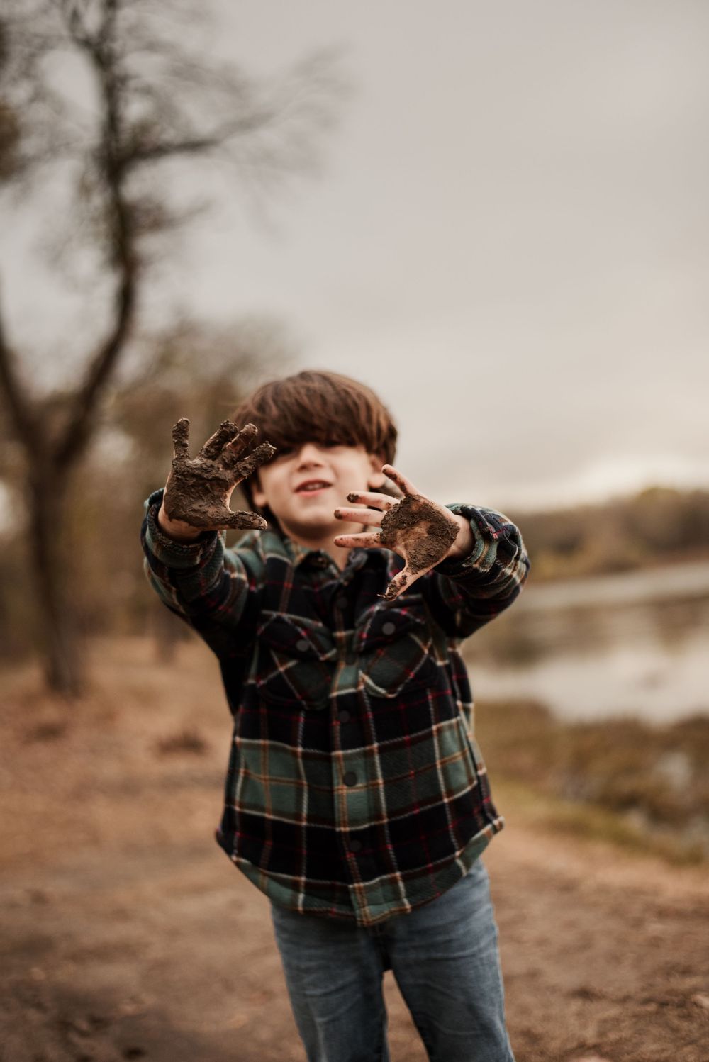 A smiling boy in a plaid shirt proudly shows his muddy hands outdoors near a river.
