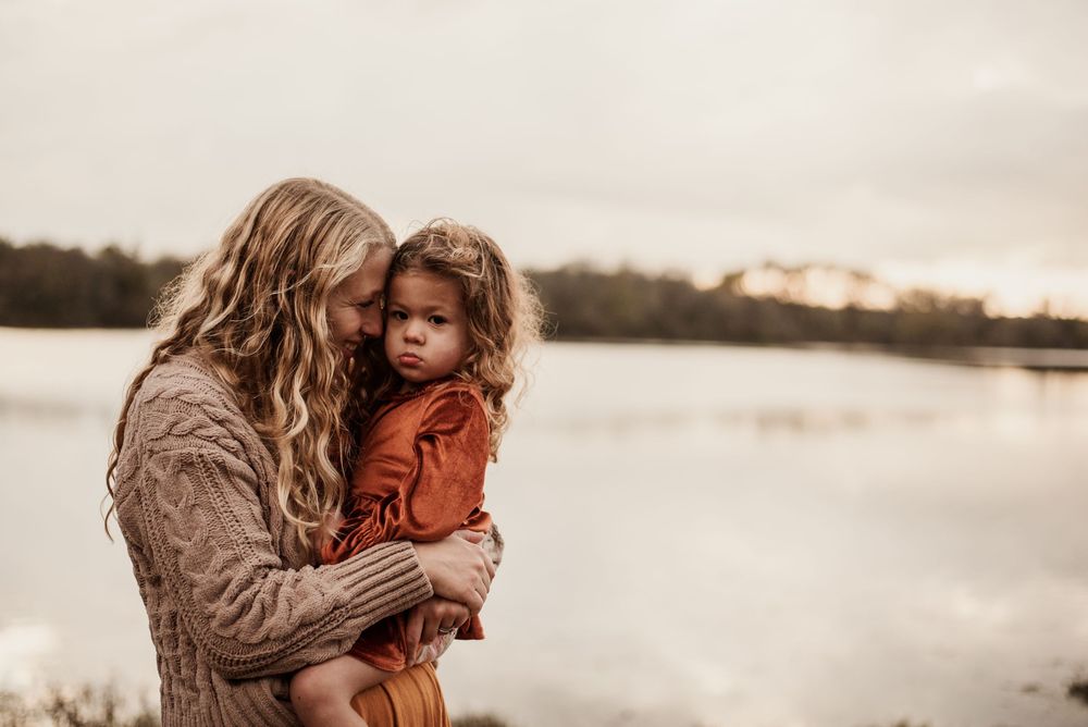 Mother with long blonde hair holds young child in rust dress near a calm lake on an overcast day.