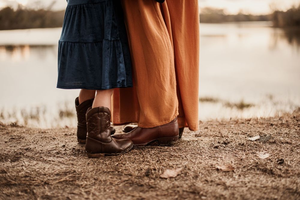 Child in cowboy boots and adult in brown leather shoes stand together near a serene lake at golden hour.
