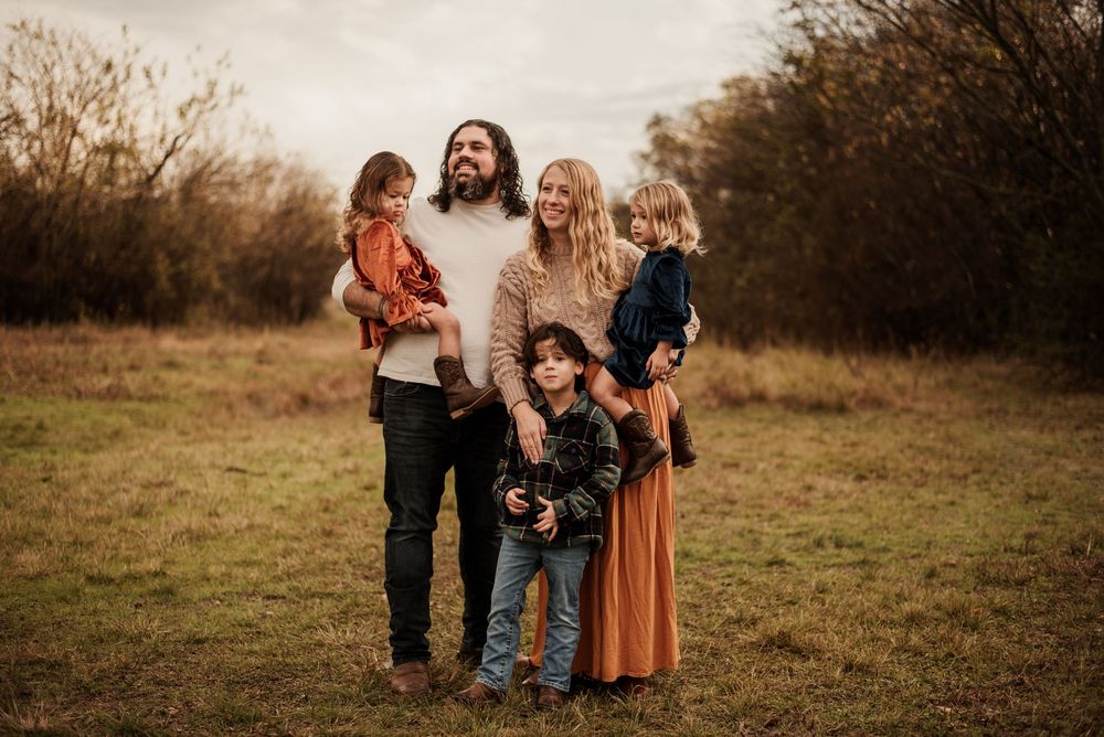 Family of five posing outdoors in a field during autumn, parents holding two young children with a boy standing in front.