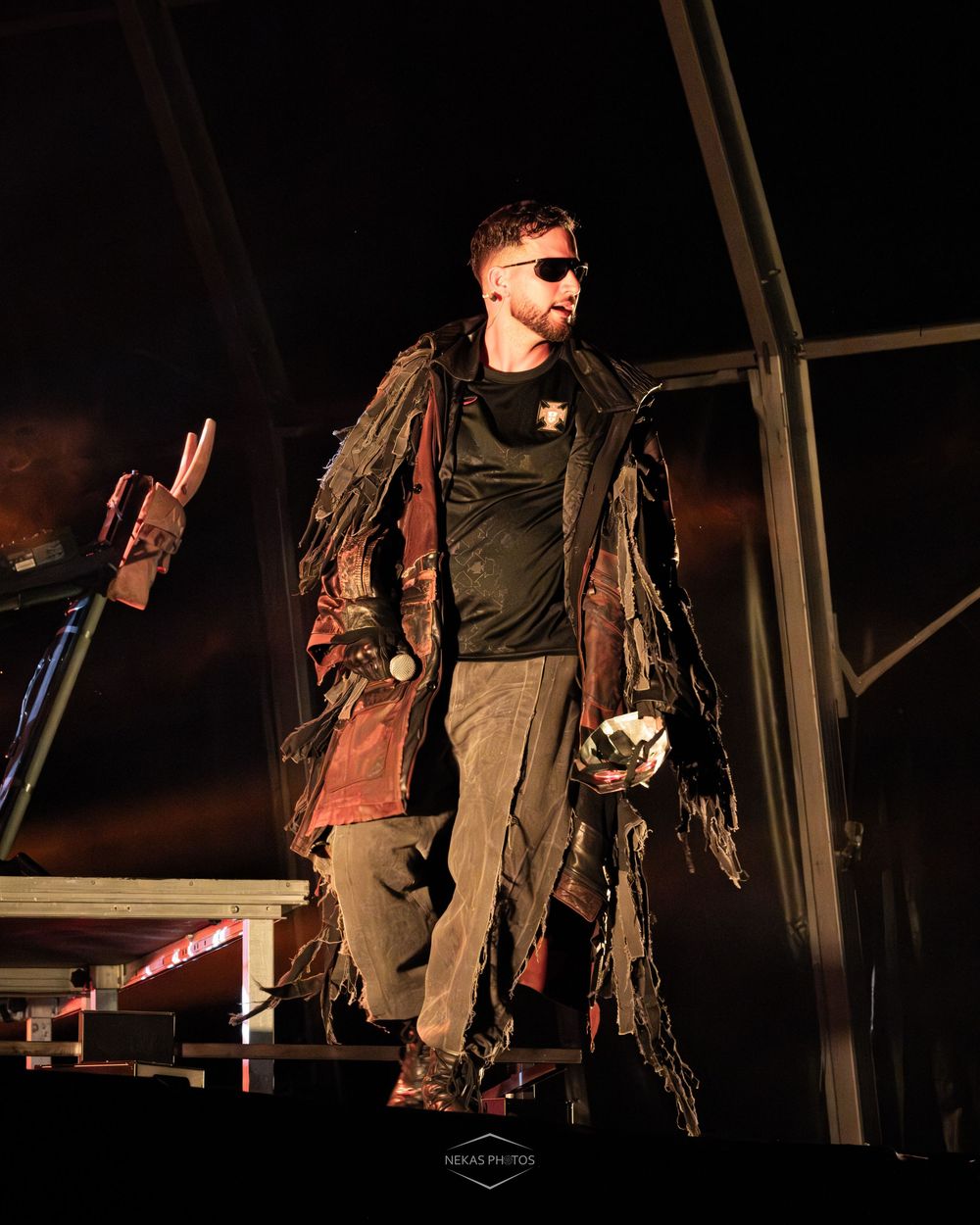 Man in tattered post-apocalyptic costume with sunglasses performing on stage at night.