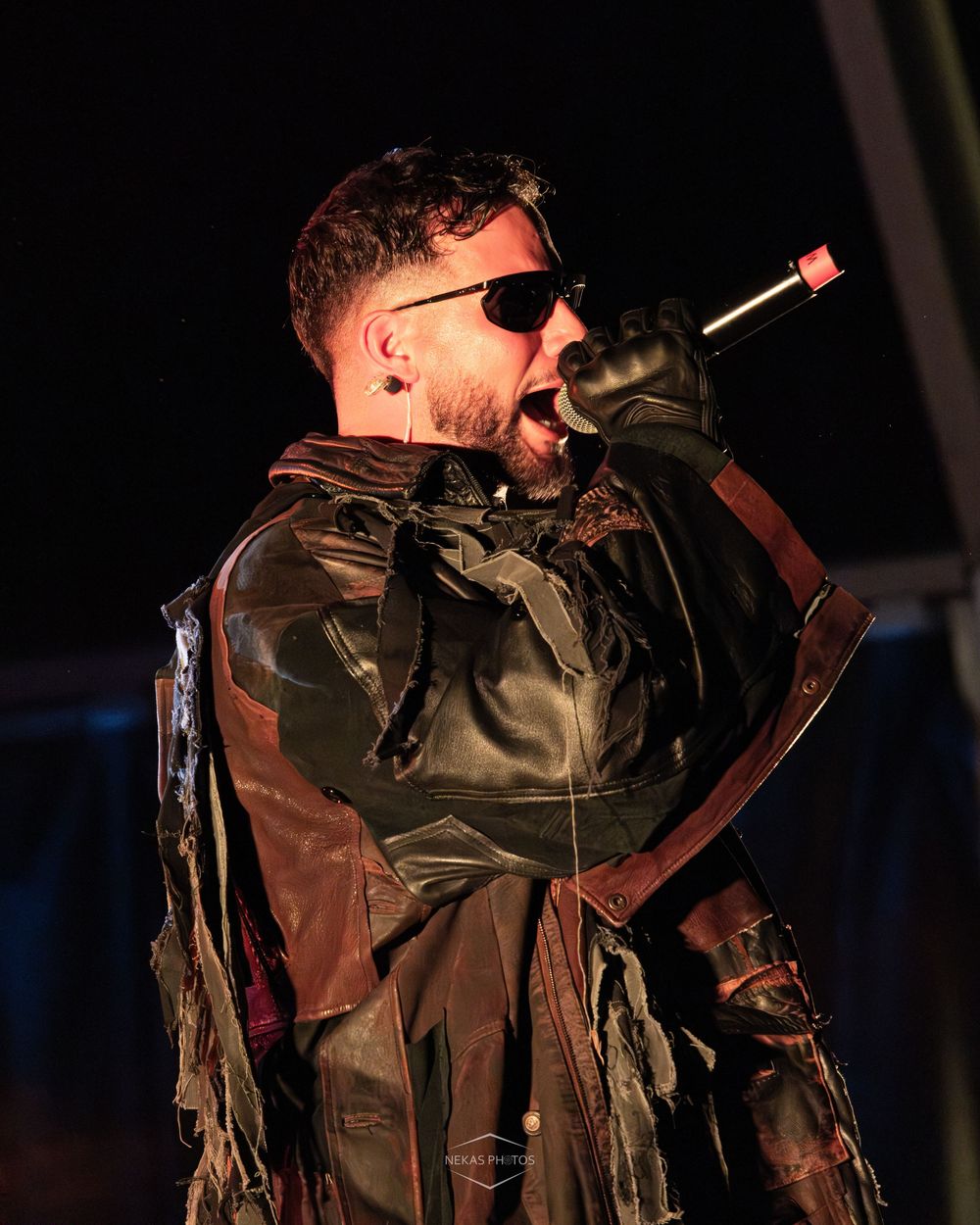 Male performer in leather costume and sunglasses singing into microphone on stage at night.