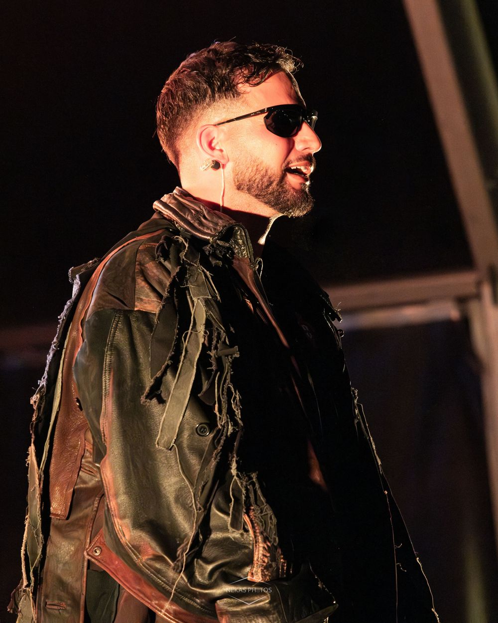 Man in black leather jacket and sunglasses with eye patch performing on stage under dramatic lighting.