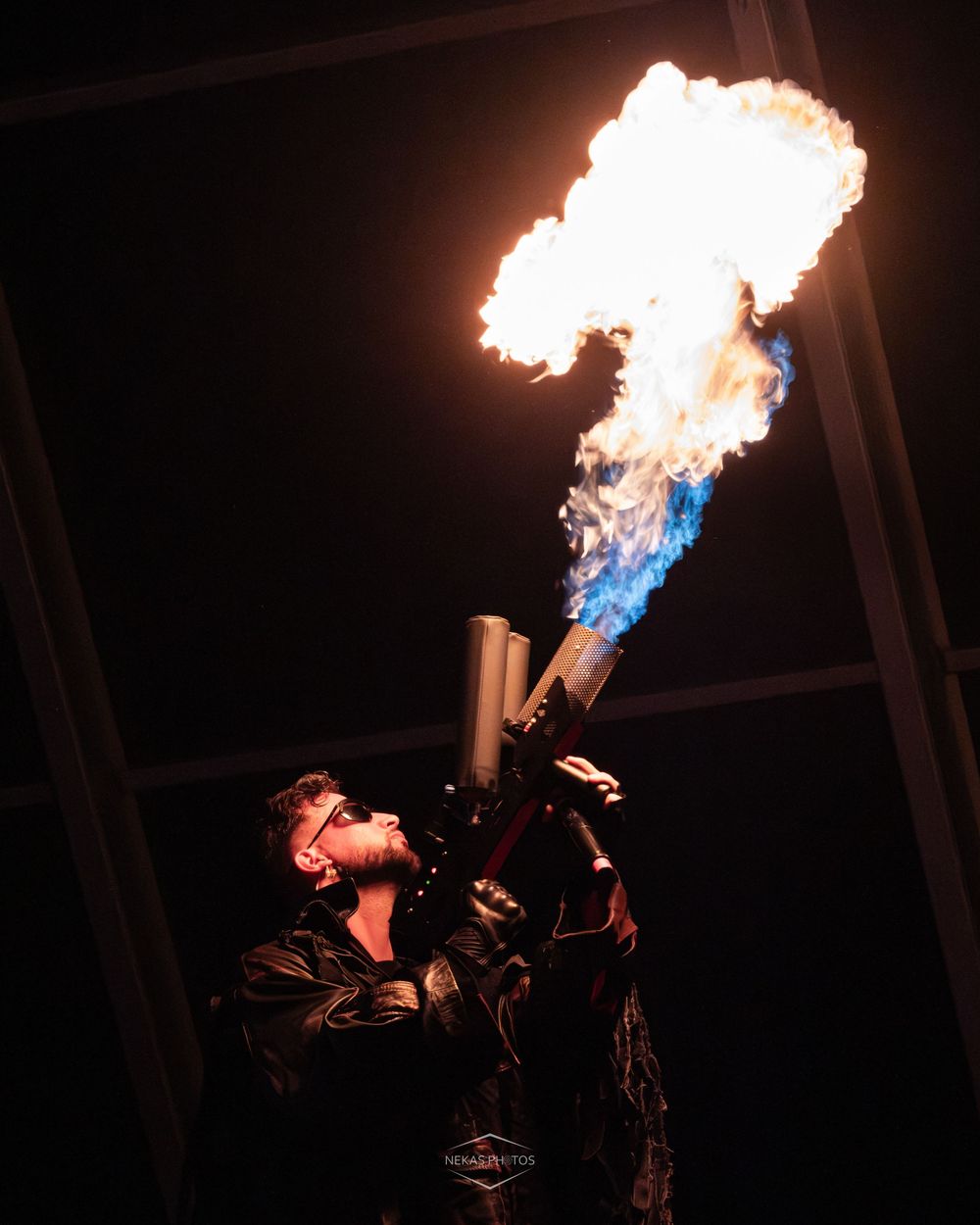 A fire performer dramatically breathes flames from a metal tube against a dark night sky backdrop.