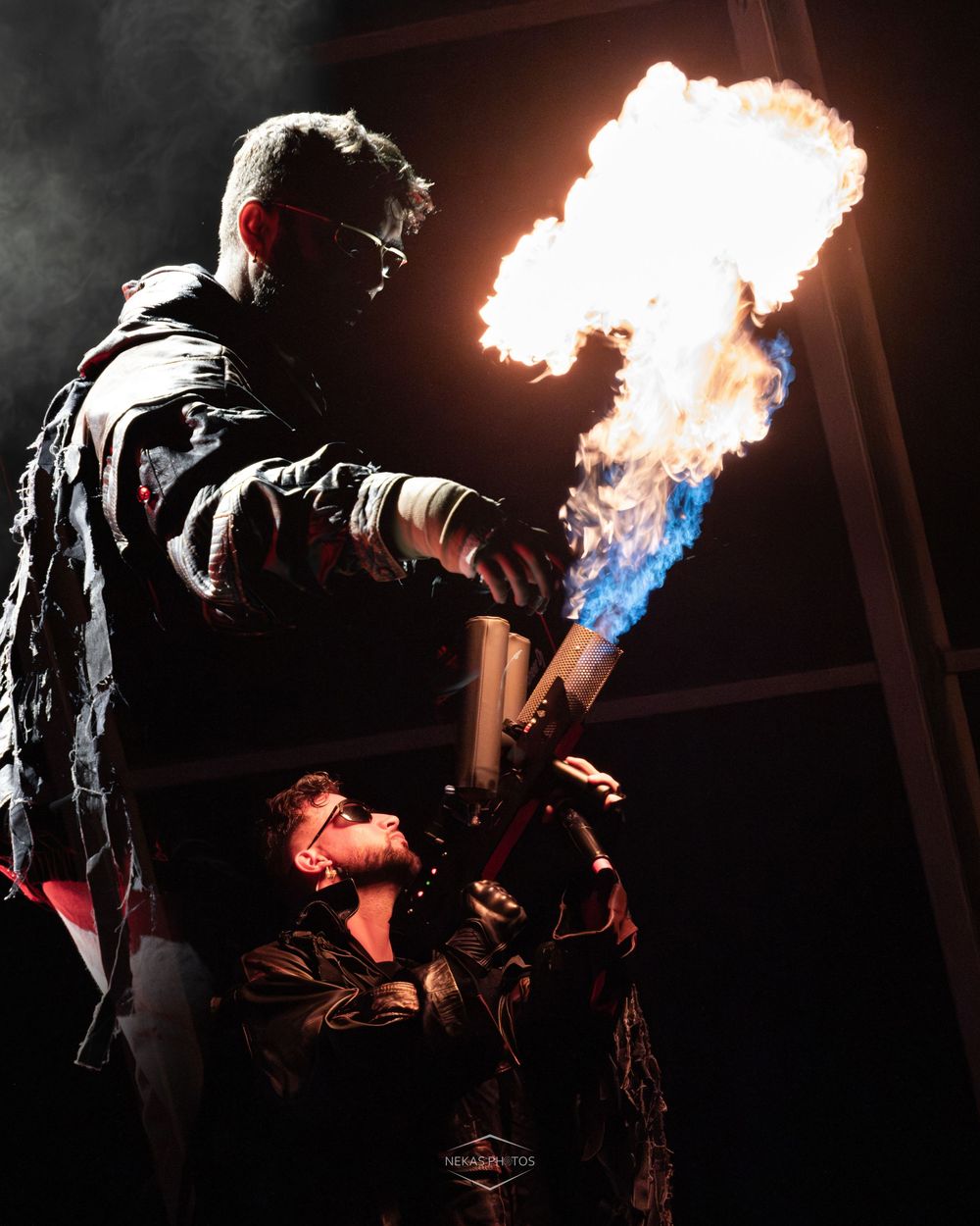 Two performers in dark jackets use a flamethrower on stage, creating a dramatic burst of fire against a black background.
