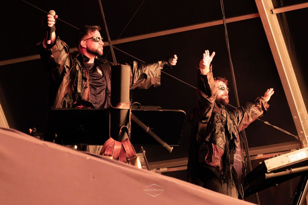 Two performers raise their hands on a dimly lit stage at night during a live music event.