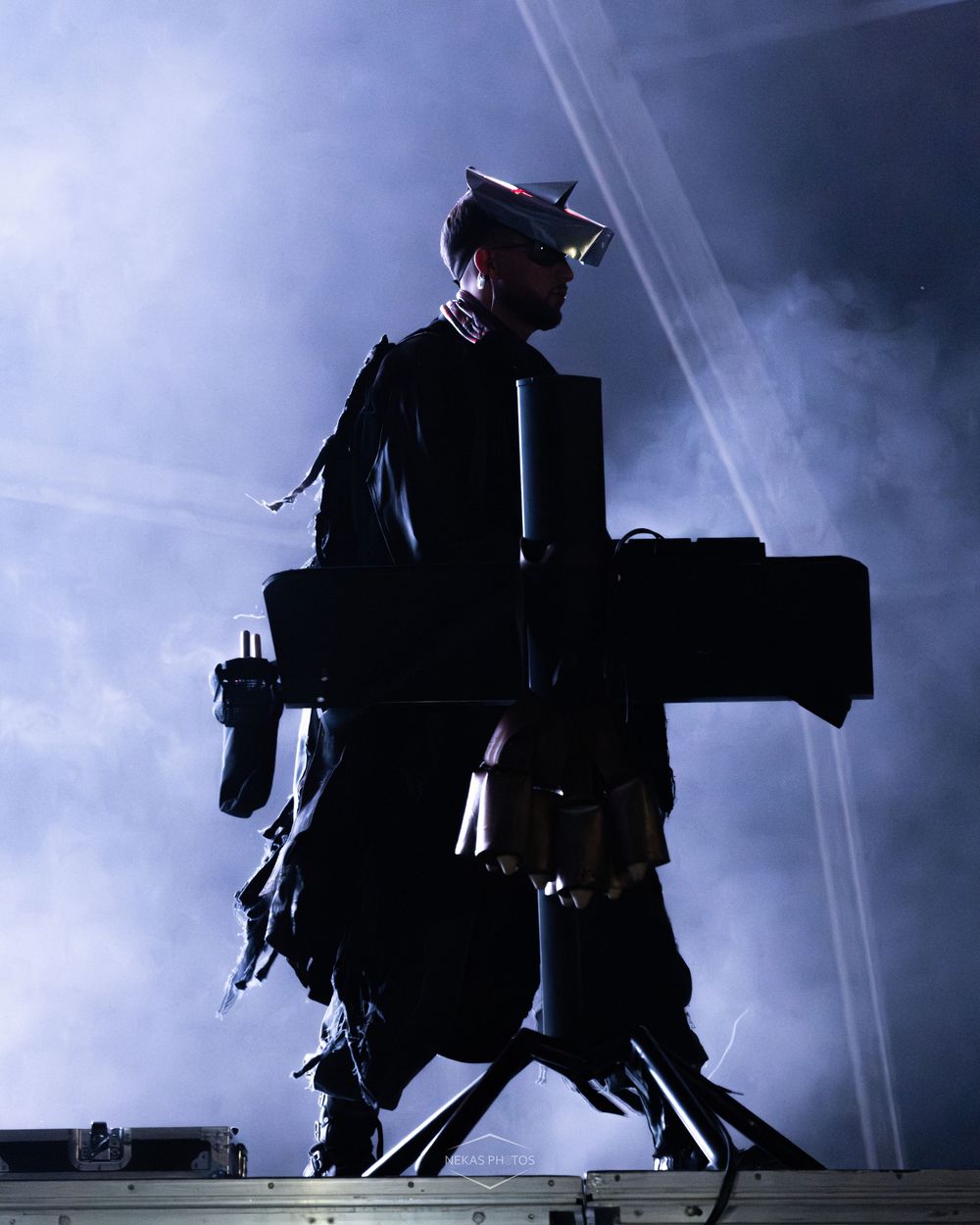 Silhouetted performer on stage wearing a military cap, surrounded by dramatic blue smoke and light beams.