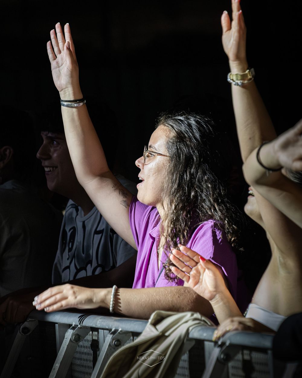 Excited woman in purple shirt raises hands at outdoor concert, wearing glasses and bracelets, crowd visible behind her.