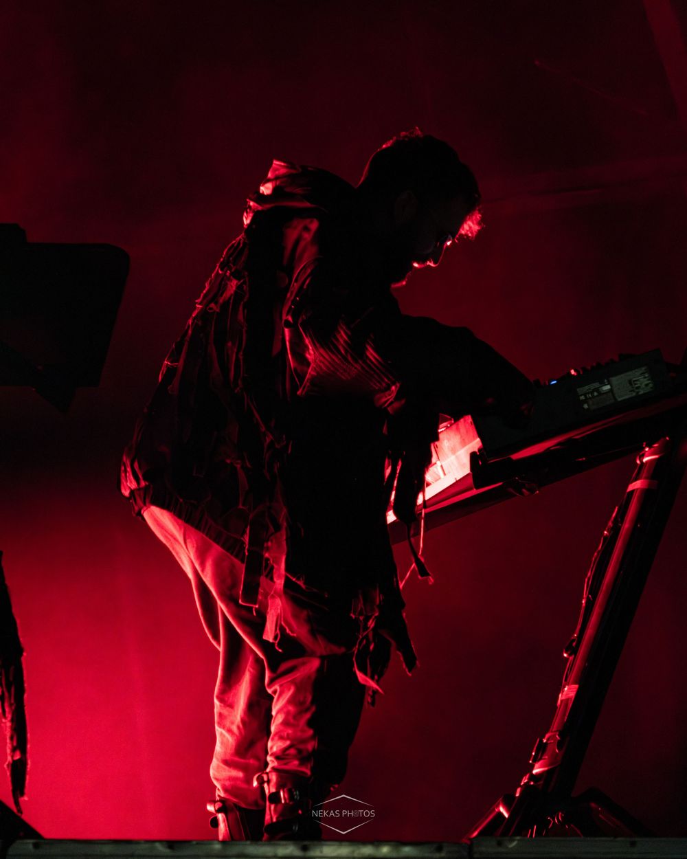 Musician performing on stage bathed in dramatic red lighting, leaning over electronic equipment during a live concert.