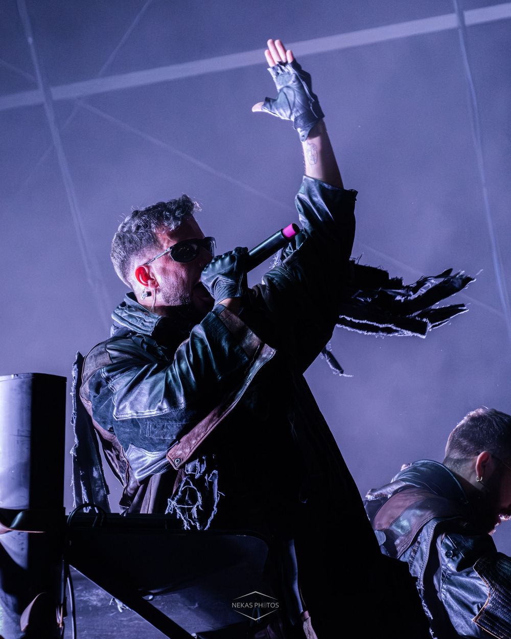A performer in a black leather jacket raises a gloved hand on a dramatic, blue-lit concert stage.