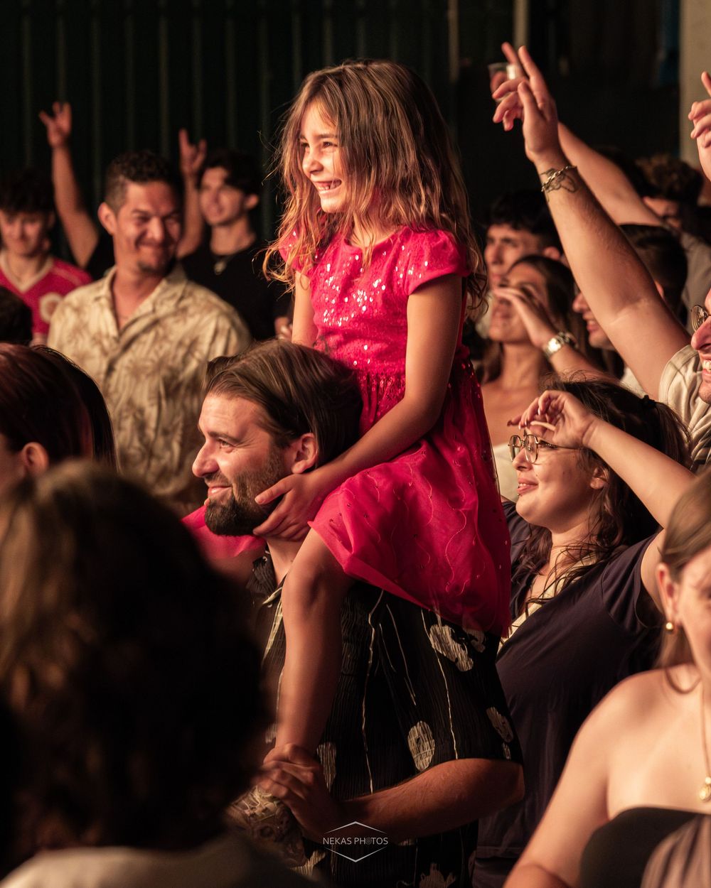 A young girl in a pink dress sits on a man's shoulders, smiling at a lively concert crowd.