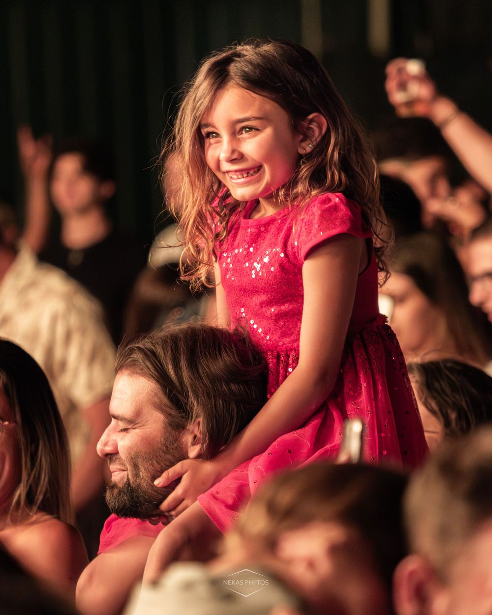 Smiling young girl in pink dress sitting on man's shoulders in concert crowd.