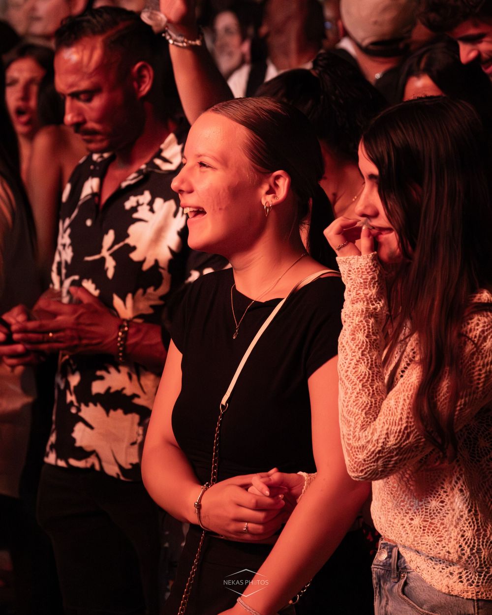 Excited crowd at a concert, woman in black top smiling joyfully, warm ambient lighting.