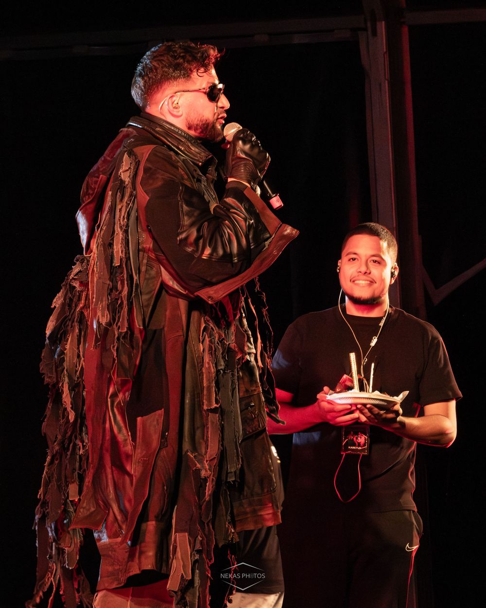 Two men on stage, one in a dramatic tattered brown coat speaking into a mic, the other smiling holding an award.