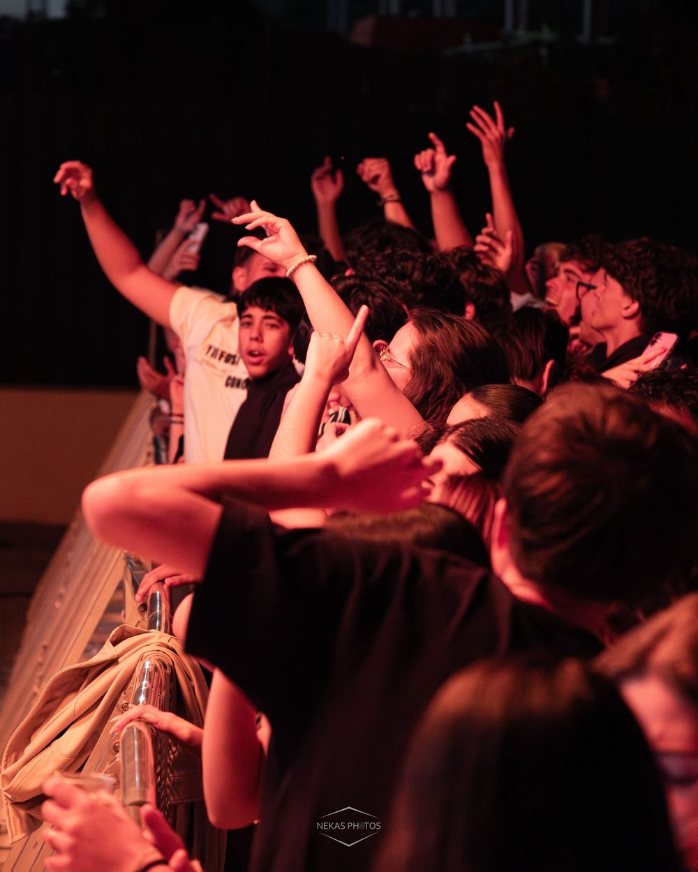 Excited concert crowd with raised hands cheering under red stage lights at a live music event.