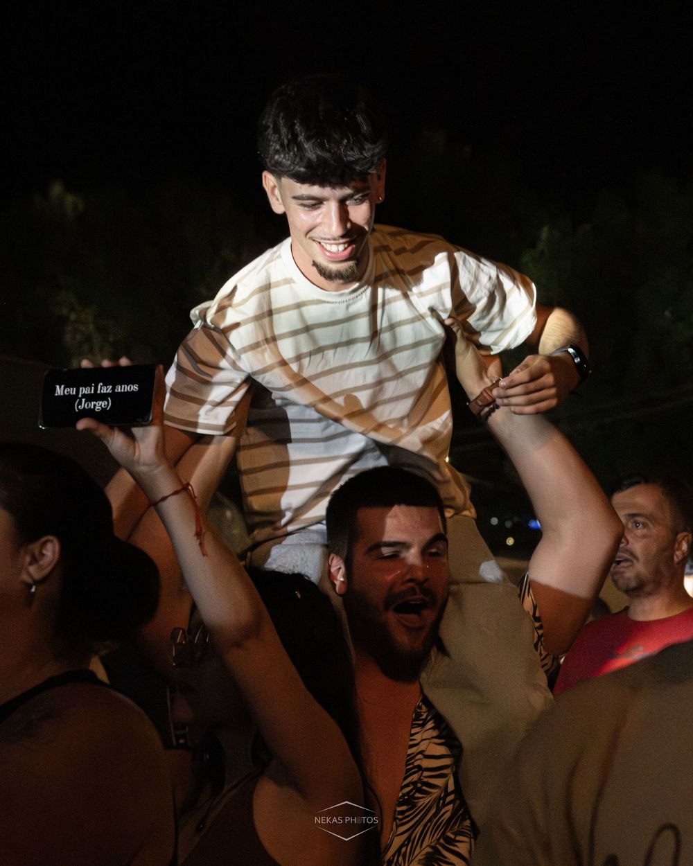 Young man sitting on someone's shoulders at a lively outdoor night party, smiling and wearing a striped shirt.