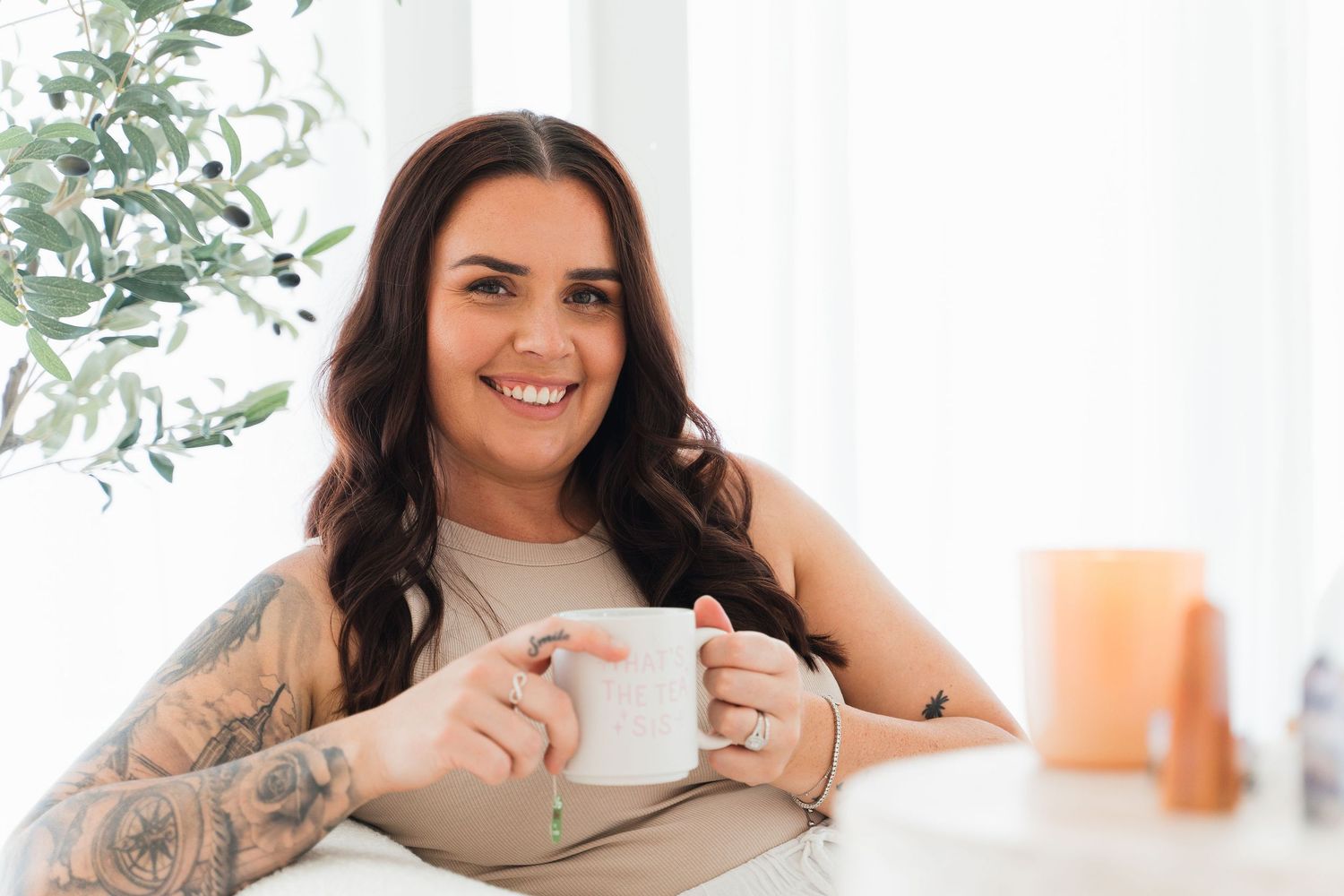 Smiling woman with tattoos holding a white coffee mug in a bright, cozy indoor setting.