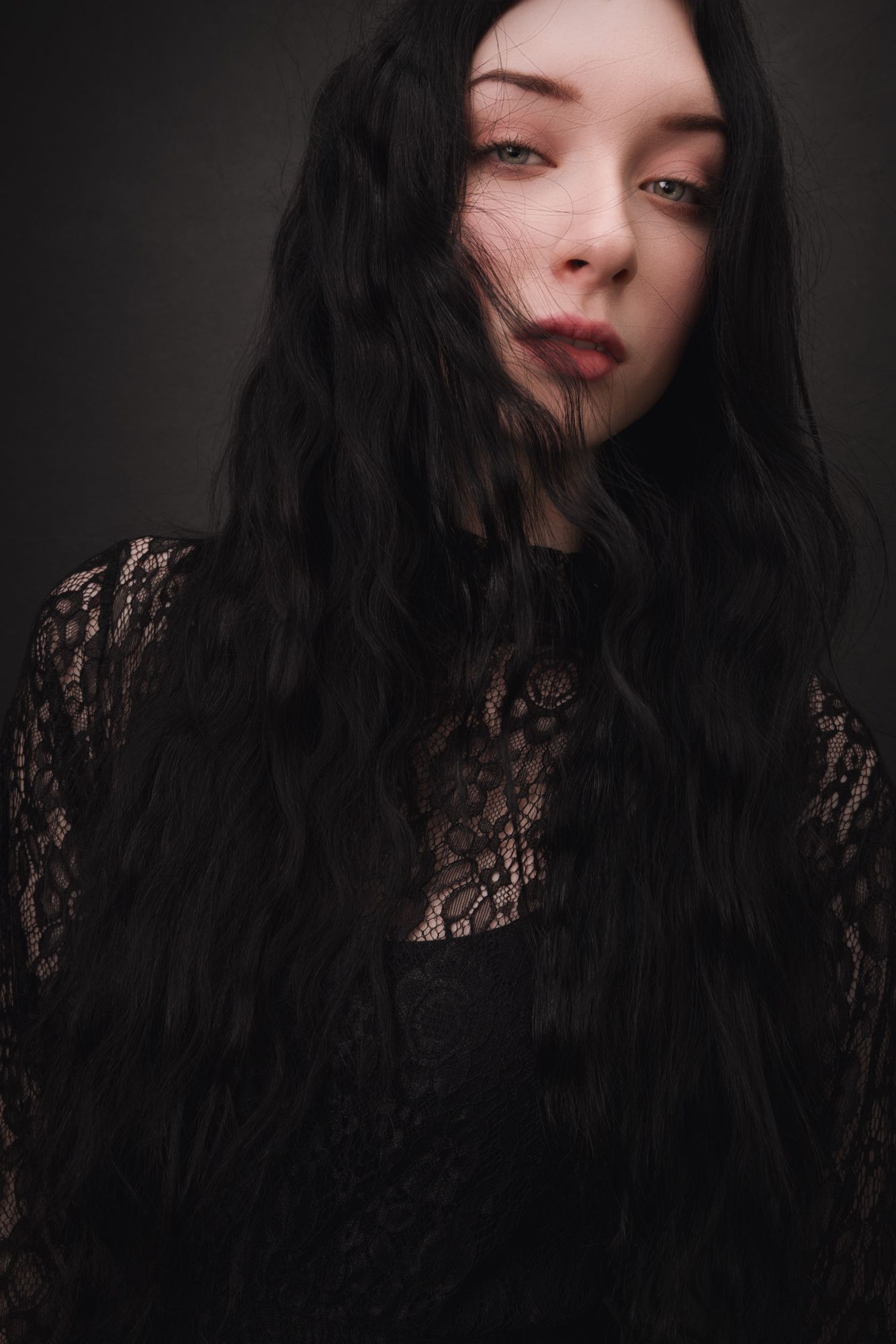 Young woman with long black wavy hair wearing a black lace top, posing against a dark background.