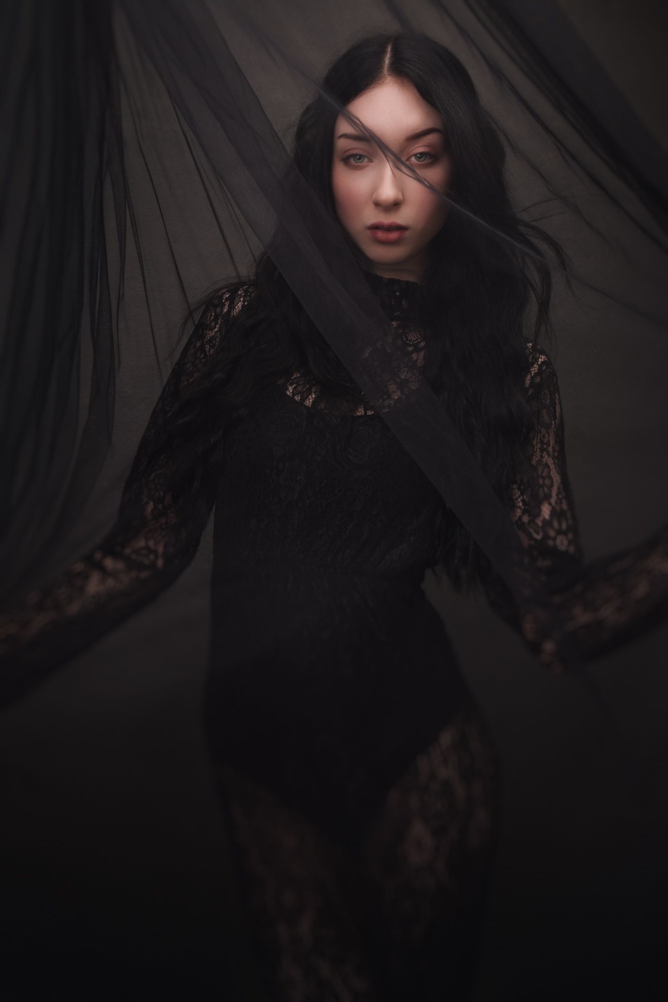 A woman in a black lace dress poses dramatically surrounded by flowing dark sheer fabric in a moody gothic portrait.