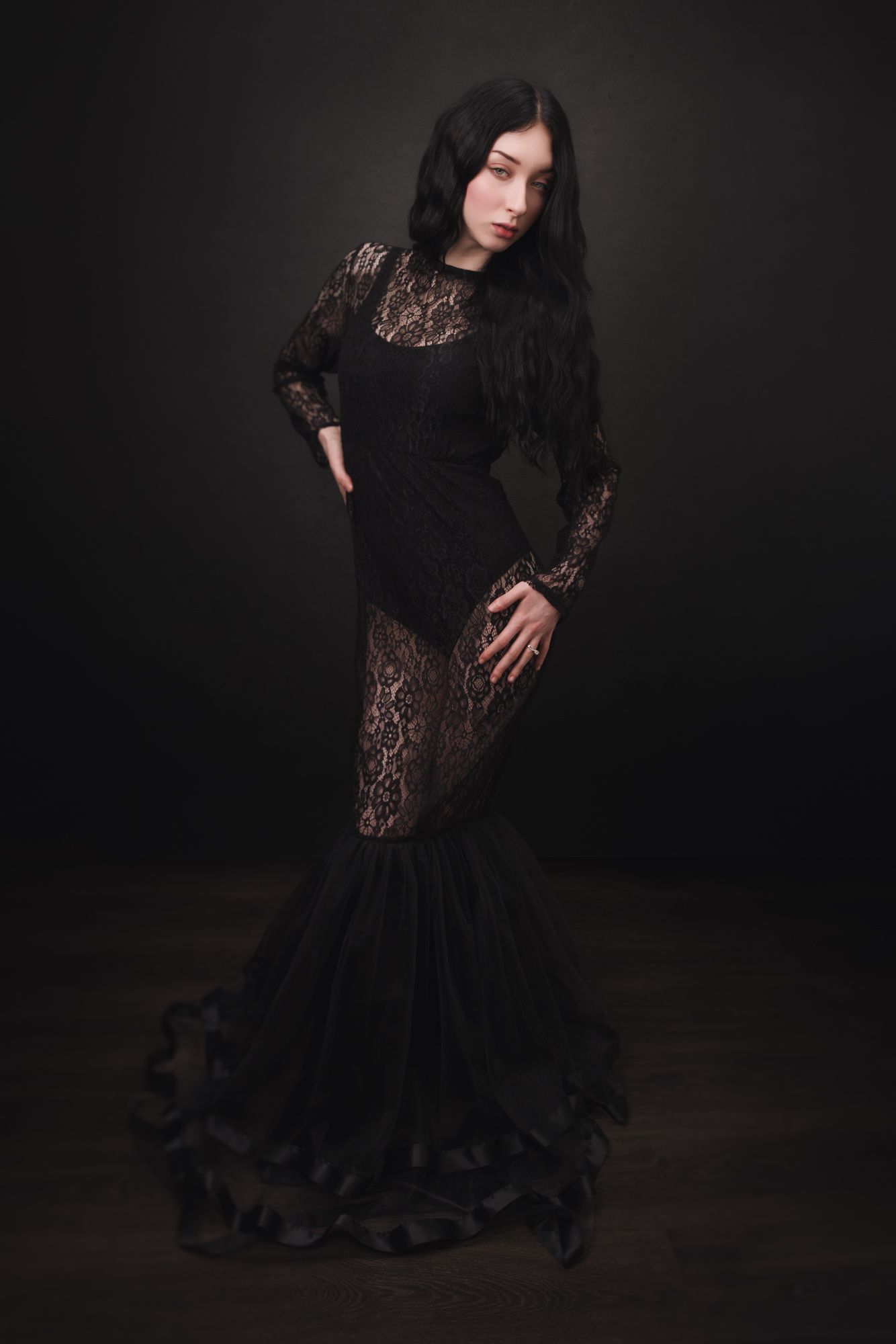 Woman in elegant black lace mermaid gown posing against dark background, long black hair, gothic fashion style.