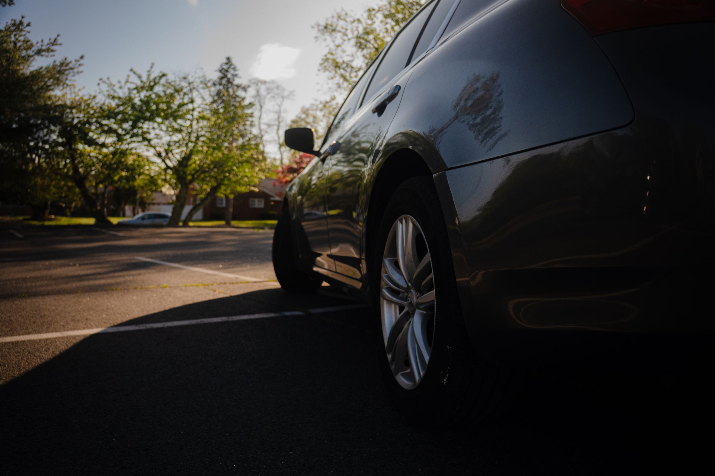 Low angle view of dark sedan car parked on sunlit asphalt lot surrounded by trees.