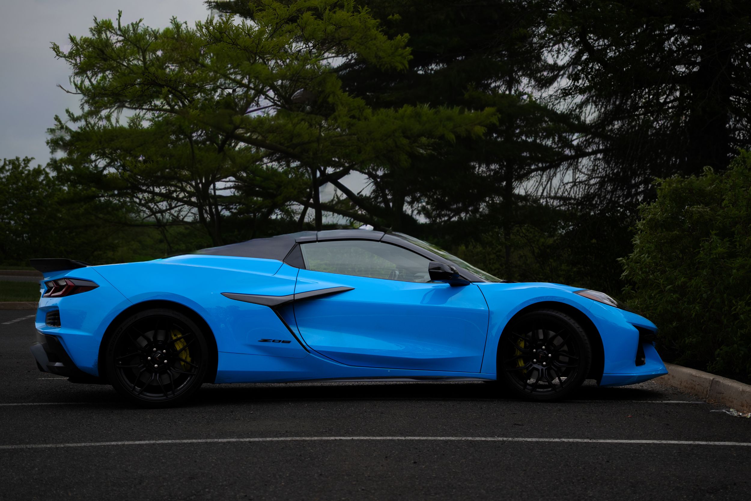Vibrant blue Chevrolet Corvette E-Ray convertible parked against lush green tree backdrop.