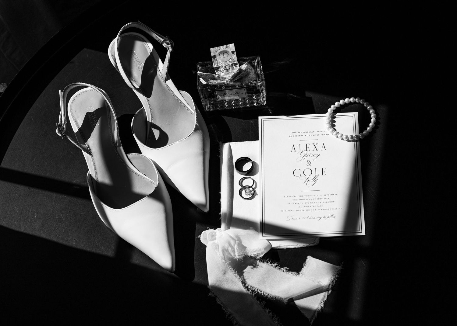 Black and white flat lay of bridal accessories including white heels, wedding invitation, pearl bracelet, and rings.