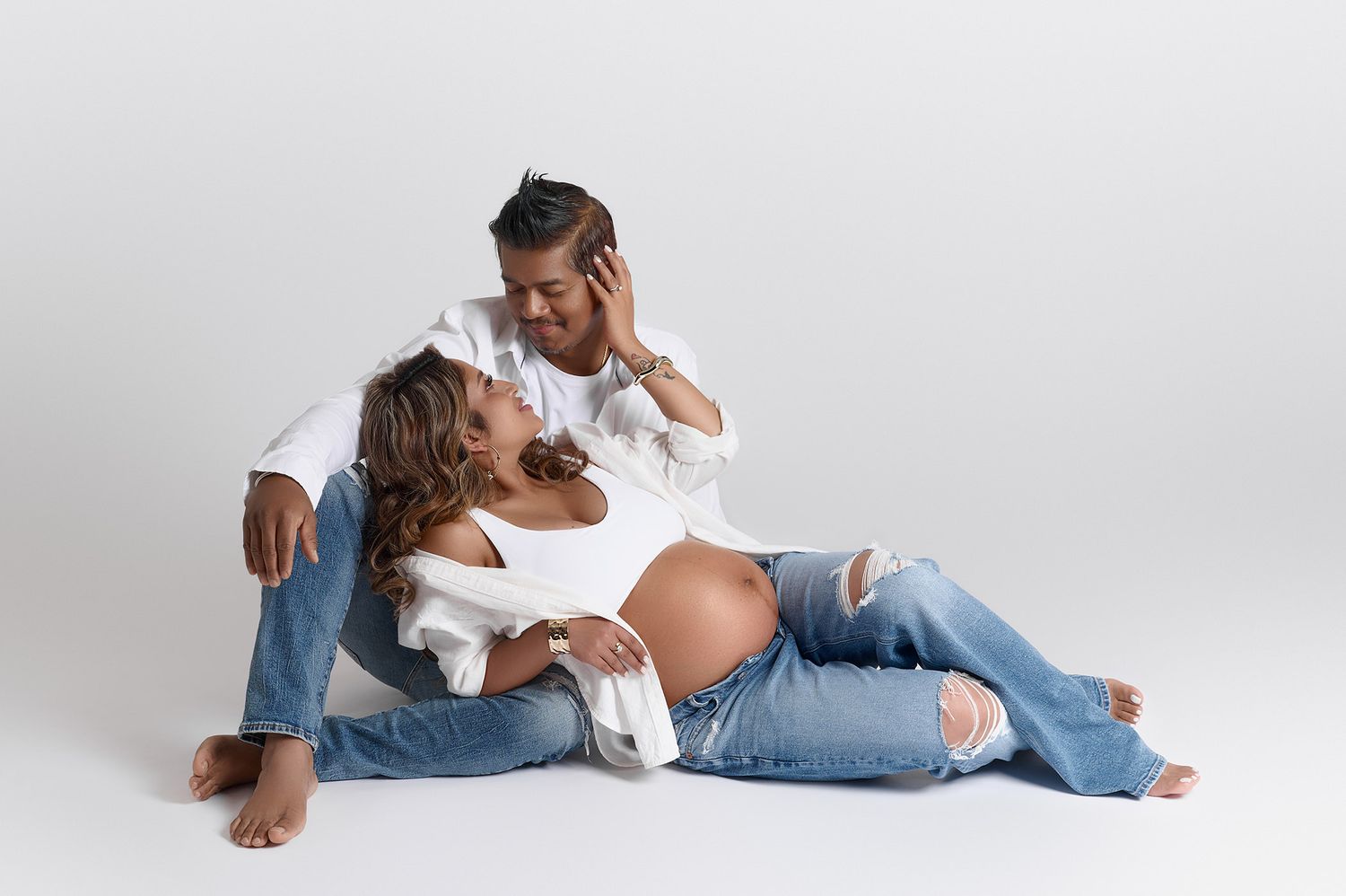 Couple in casual white and denim outfits posing together for maternity studio portrait.