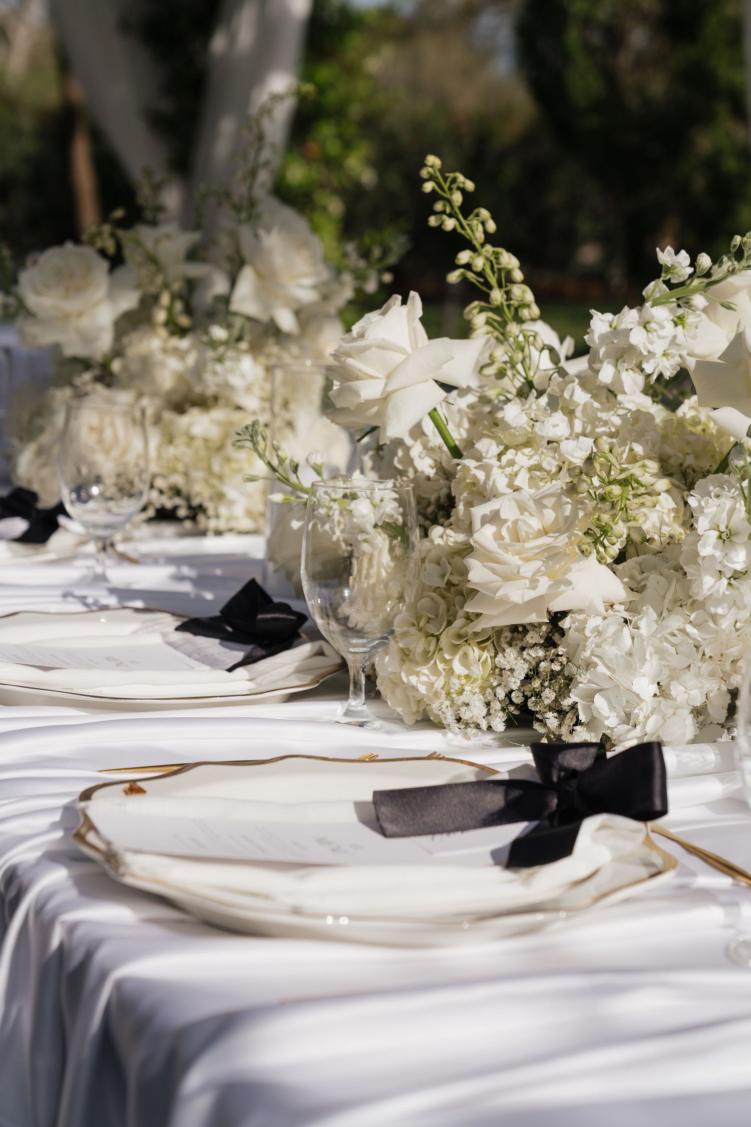 Elegant outdoor wedding table setting with white floral centerpieces, black bow napkins, and white plates on a satin tablecloth.